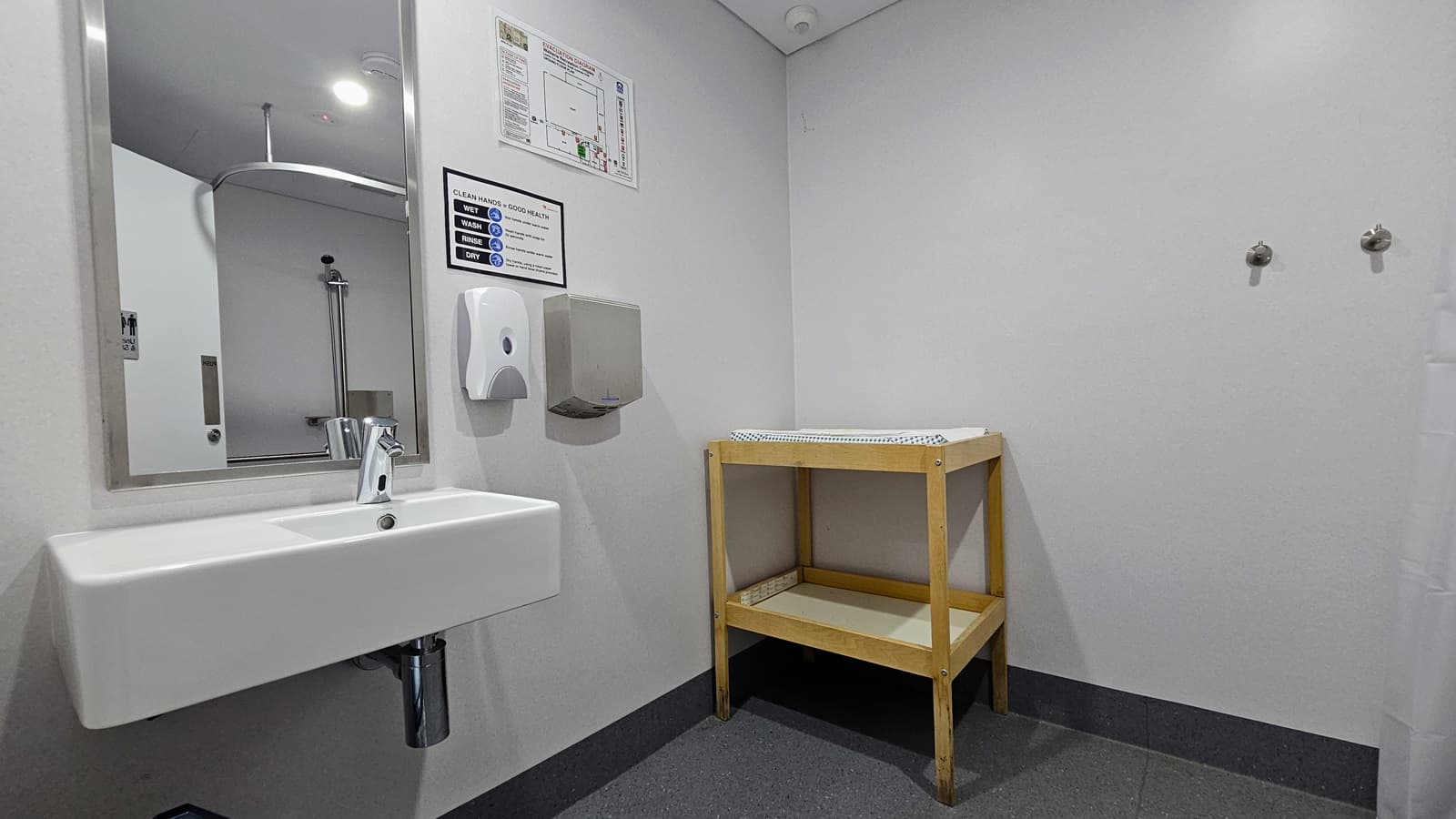Inside the bathroom, a two-layer wooden table is positioned in the corner. On the left, a washbasin, a glass mirror, a liquid soap, and a tissue dispenser are mounted on the wall, along with banners displaying the site location and safety reminders. The floor is grey, and the walls and ceiling are both white.