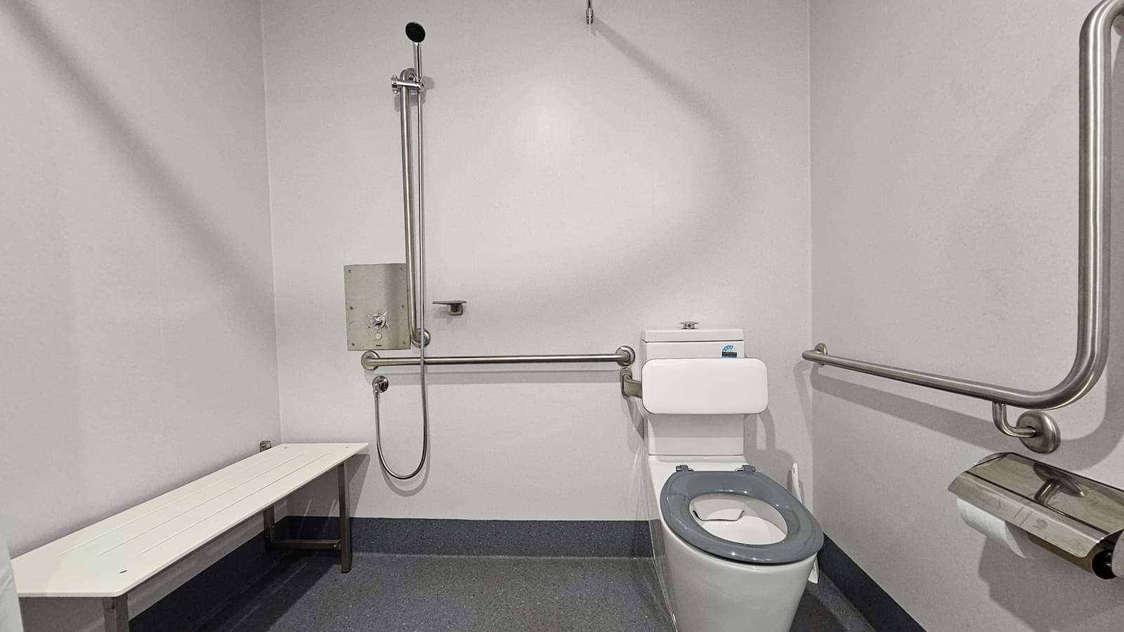 A closer look at the bathroom reveals a long white bench positioned on the left wall. In the middle, there are showers equipped with handrails. Beside them is a toilet bowl also equipped with handrails. A tissue dispenser is mounted to the wall on the right. The walls are all white, and the floor has grey tiles with a granite design.