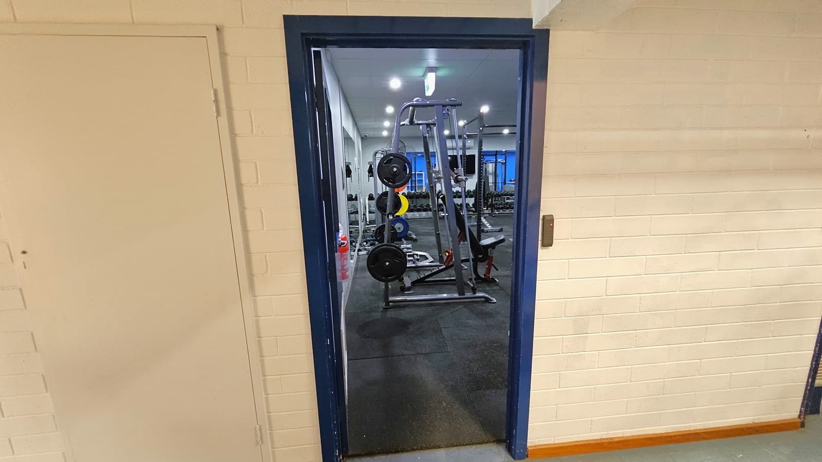 On the left is the reception next to a blue door. A free standing banner stands in the middle, accompanied by framed posters and a fire hose reel. To the right, a corridor leads to the Stadium, with pamphlets placed on the wall. A staircase with a ramp leads upstairs. In the corridor, there's a staircase in the middle, a fire exit sign, and posters on the left wall. The ceiling transitions from white outside to cream with a corrugated design in the corridor. A rubbish bin is placed on the right.
