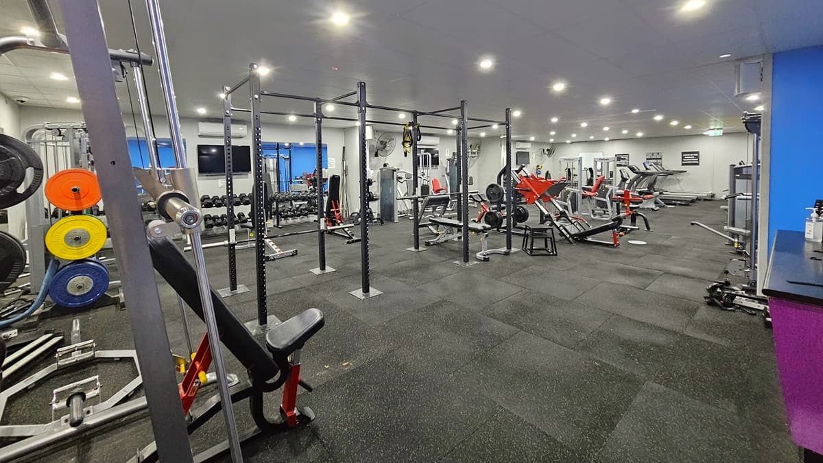 To the right is a fuchsia counter table with hand sanitizers. On the left, weightlifting machines, lat pulldown machines, and smith machines are arranged. In the middle row, there are rowers, bench machines, and treadmills. Further in, the weights equipment, including barbells and kettlebells, are arranged on the left. A flat-screen TV is mounted on the left wall. The wall is white with blue accents, and the floor has grey rubber tiles.