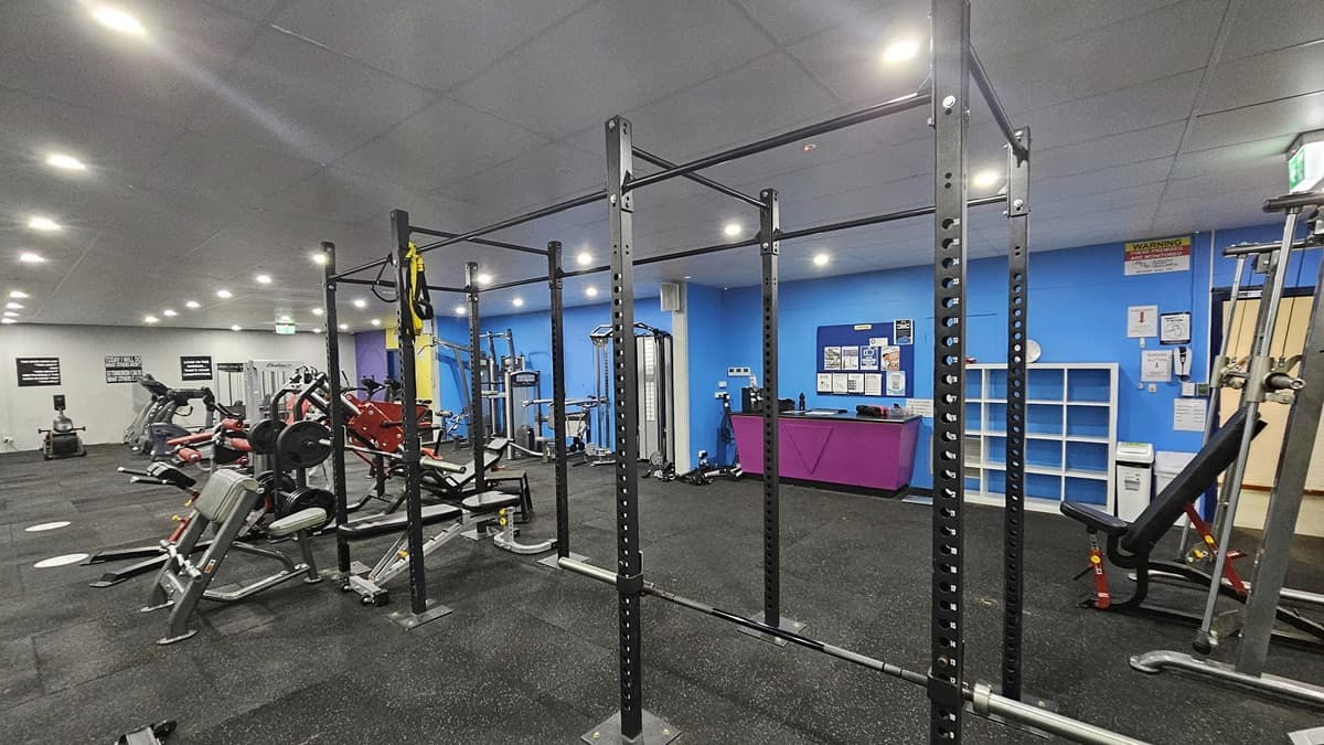 A fuchsia counter table with hand sanitizers is placed on the middle wall, accompanied by a display rack for belongings. To the right is a lat pulldown machine, and in the middle row, a smith machine is positioned along with rowers, bench machines, and treadmills. Further in, additional machines are arranged. The ceiling is entirely white with a porous design, and the floor is covered in grey rubber tiles. Posters adorn the blue wall on the right, and the left wall is white.