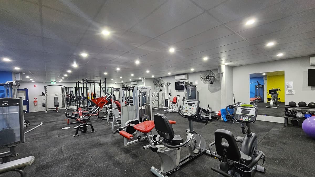 To the left is a bench press machine, and in the middle row are a lat pulldown machine, smith machine, rowers, bench machines, treadmills, and other machines. Further in, additional machines are arranged. The ceiling is entirely white with a porous design, and the floor is covered in grey rubber tiles. Flat-screen TVs are mounted to the right wall, along with wall fans. A floor-to-ceiling mirror is mounted to the right wall and adjacent are the weights and stability balls.