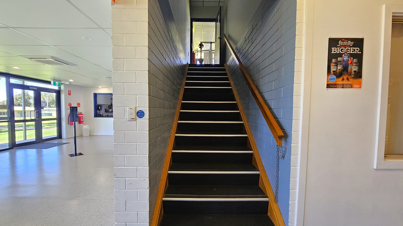The stairs leading to the second gym are equipped with wooden handrails, and a chain is hanging at the tip. A poster is placed on the right wall, which is made of white bricks. On the left, the glass door serves as the entrance to the building. In the middle stands a hand sanitizer dispenser, and a section of the reception is also visible.
