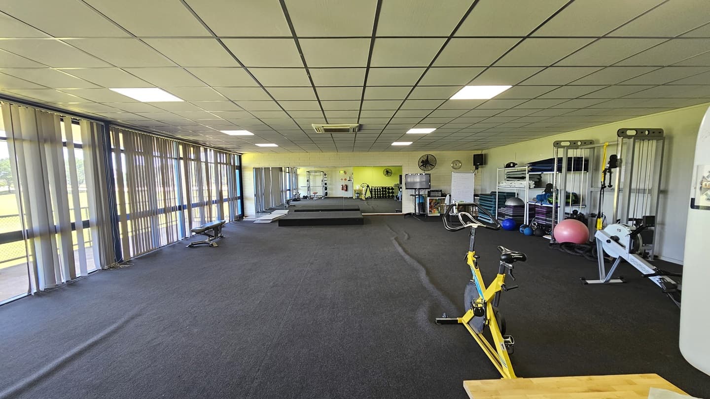 Machines, including a Smith machine and a rower, are lined up on the right. A stationary bike is placed in the middle, and mats, stability balls, and medicine balls are on the right. In the center are two elevated platforms. On the left, a bench machine is positioned near the windows, which have floor-to-ceiling glass partially covered by vertical blinds. Affixed to the right wall are a flat-screen TV, clock, audio speaker, and wall fans. The ceiling is white, and the floor is carpeted in grey.