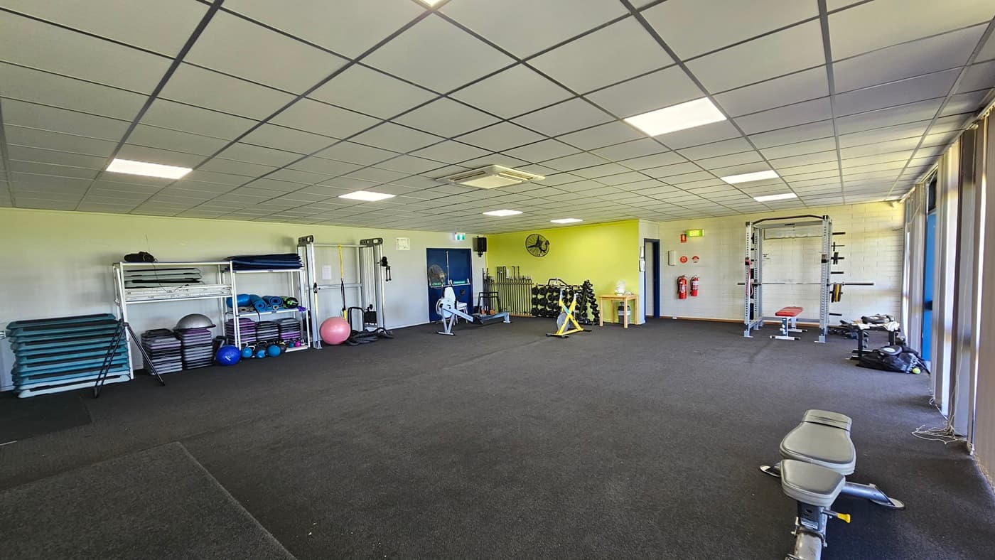Machines, including a Smith machine and a rower, are lined up on the left. Mats, stability balls, and medicine balls are also on the left. In the corner, a flat-screen TV and a wall fan are affixed. On the right, a bench machine is positioned near the windows, which have floor-to-ceiling glass partially covered by vertical blinds. Another Smith machine is placed near the wall. Two fire extinguishers are affixed to the middle wall. The ceiling is white, and the floor is carpeted in grey.
