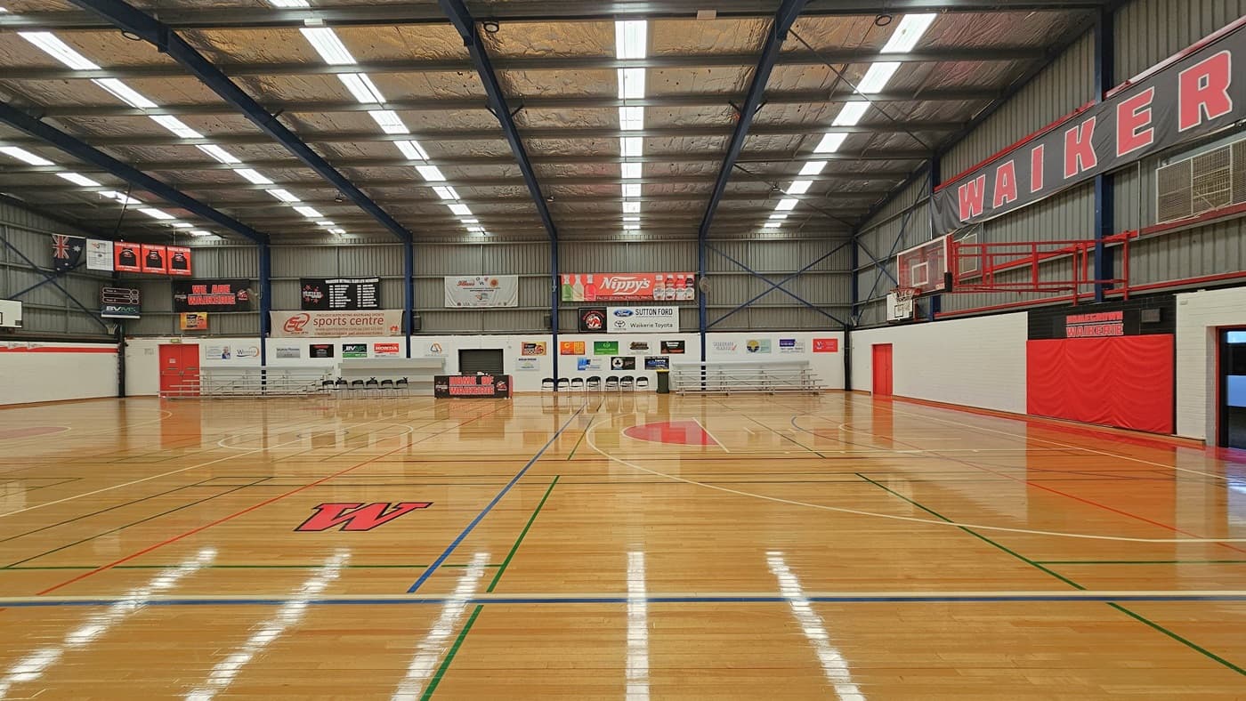 On the right, a white brick wall hosts a defibrillator in a red wall-mounted box with informational instructions. The left wall holds three picture frames and light switches. A short aluminum ramp covers an approximately 10cm stepped entry to the stadium. Inside, the stadium features highly varnished timber flooring with markings for various ball games, and tiered seating is arranged on both sides. The ceiling exhibits exposed blue beams, and fluorescent lights. Banners encircle the area.