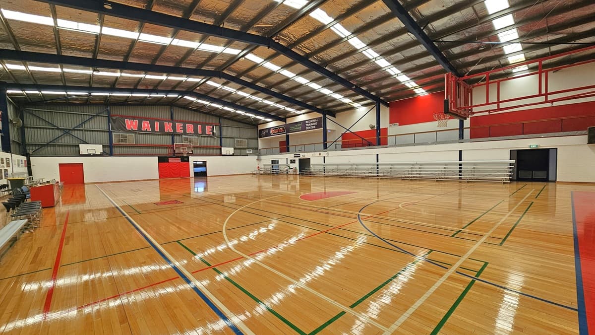 In the stadium, the floor is polished timber marked for various ball games. The ceiling has insulated foams, exposed blue beams, and fluorescent lights. Black and red banners displaying the local Waikerie basketball team encircle the area. Red doors are on the left, and another door is open on the right. A white bench and black plastic chairs are aligned on the left, while tiered seating is arranged on the right.