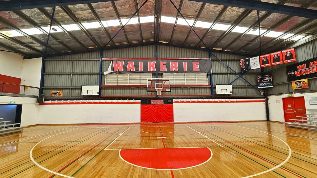 In the stadium, the floor is polished timber marked for various ball games. The pitched ceiling features insulated foams, exposed blue beams, and fluorescent lights. Black and red banners displaying the local Waikerie basketball team encircle the area. Red doors are situated on the right, with tiered seating on both sides of the court. A digital screen is affixed to the upper right-hand corner, and an audio speaker is positioned on the left.