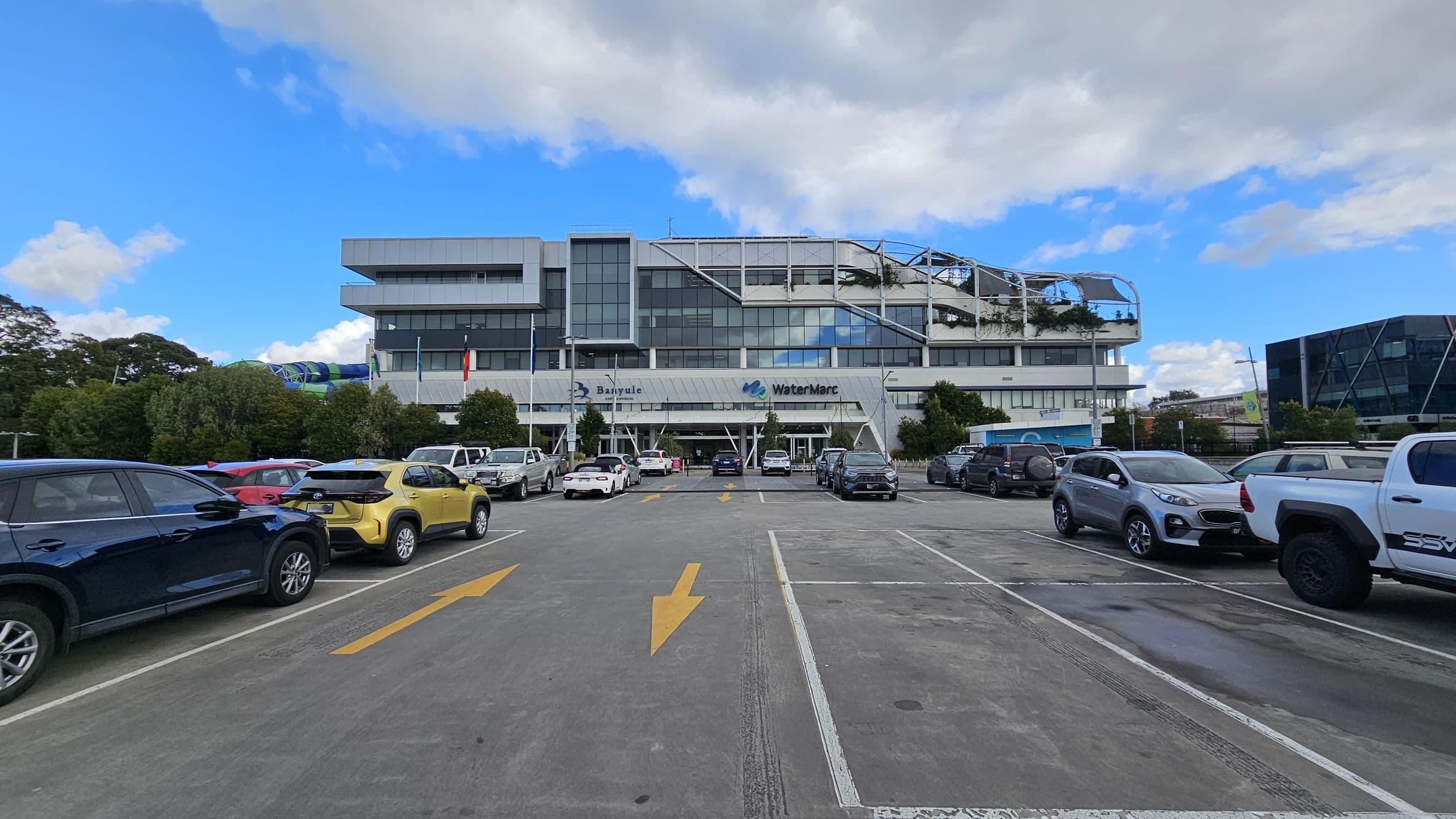A large, modern building with a glass facade is in the centre, featuring the names "Banyule" and "WaterMarc" on the front. The foreground shows a car park with various vehicles, including a yellow car and a white truck. Yellow directional arrows are painted on the concrete surface. Trees and bushes line the perimeter of the car park, and another building with a geometric design is visible on the right.