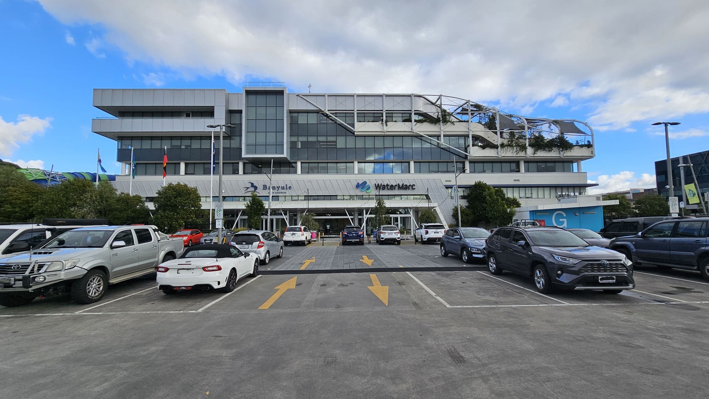 A multi-storey building with a modern design, featuring large glass windows and a rooftop garden with greenery. The building has signs for "Banyule" and "WaterMarc" on the front. In the foreground, a car park with various vehicles, including a white convertible and a silver pickup truck. Yellow arrows are painted on the asphalt, indicating traffic direction. Trees and flagpoles are positioned near the entrance. The sky is partly cloudy.