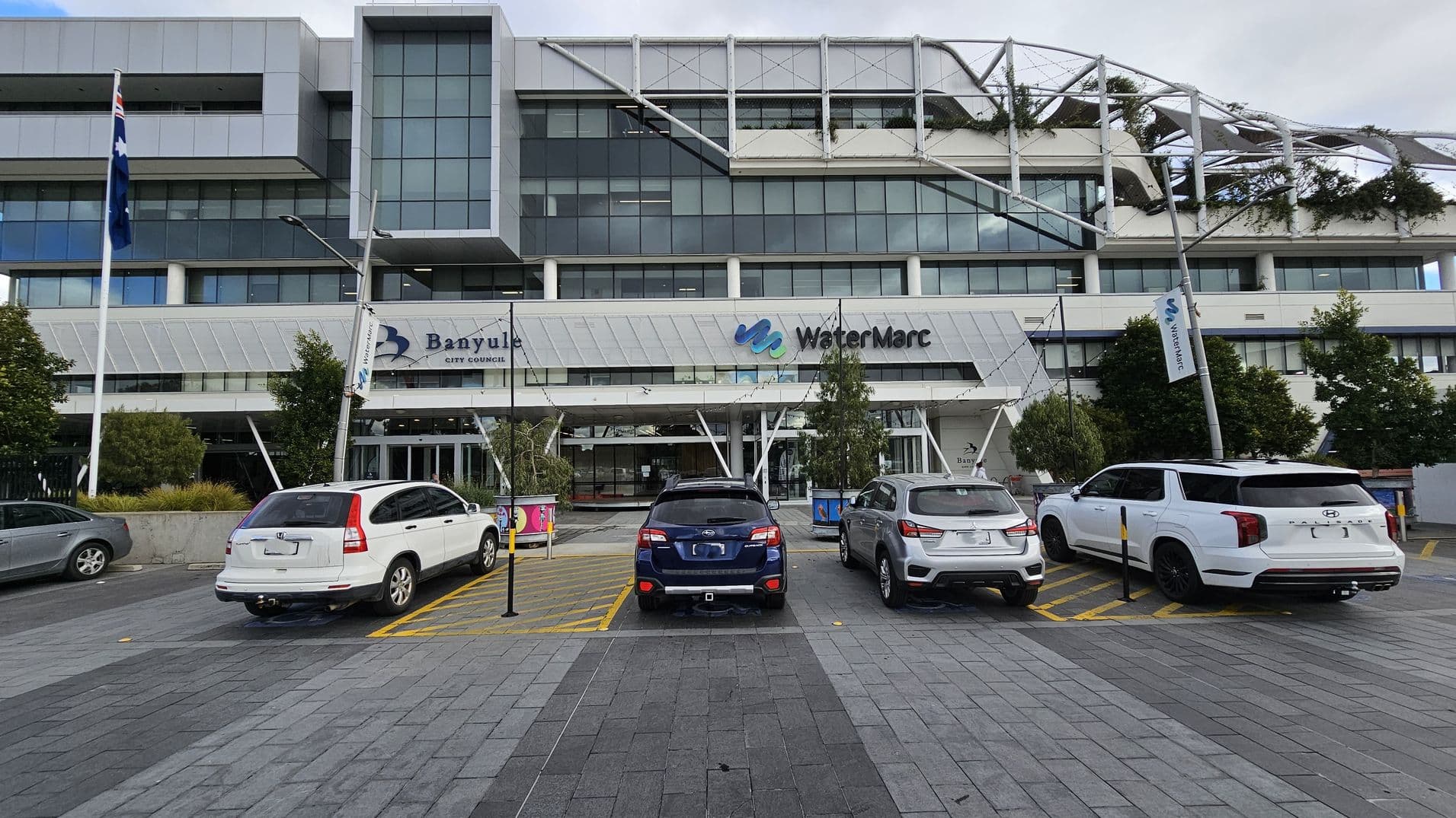 A modern multi-storey building with large glass windows and a geometric design. In the foreground, there are four parked cars on a paved area. The building features signage for "Banyule City Council" and "WaterMarc" above the entrance. There are several trees and a flagpole with a flag on the left. The structure has a distinctive roof with greenery and metal framework. The pavement is grey with a pattern of rectangular tiles.