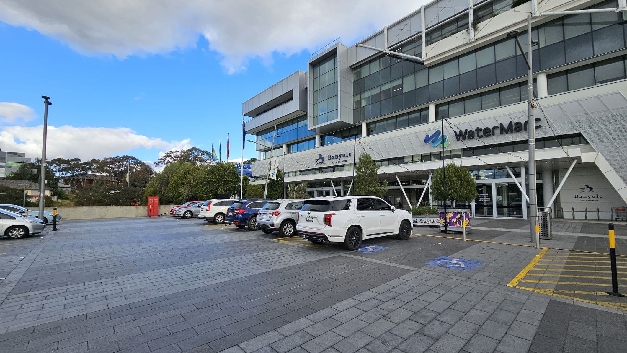 A modern building with large glass windows and a sign reading "WaterMarc" and "Banyule City Council" on the facade. Several cars are parked in the foreground on a paved area. Flags are visible near the entrance, and there are trees and bushes along the side. A red utility box is positioned near the centre-left. The building has a sleek, angular design with metal and glass elements.