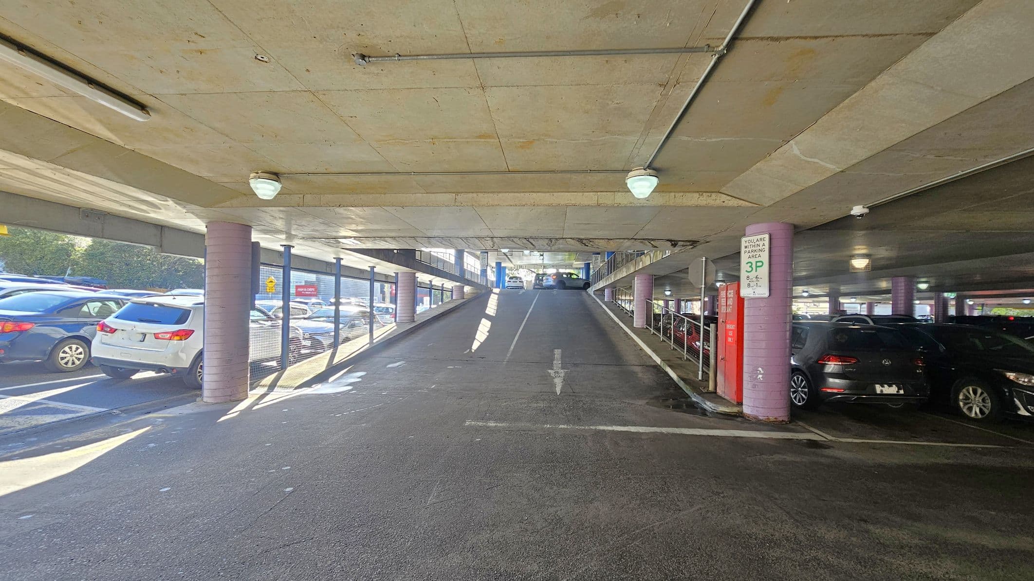 A multi-storey car park with a ramp in the centre leading upwards. On the left, several parked cars are visible, including a white SUV. On the right, more cars are parked, with a sign indicating parking restrictions. The ceiling is concrete with lights, and the floor is asphalt. Pink cylindrical columns support the structure, and metal railings line the ramp. Natural light filters in from the left side, illuminating the area.