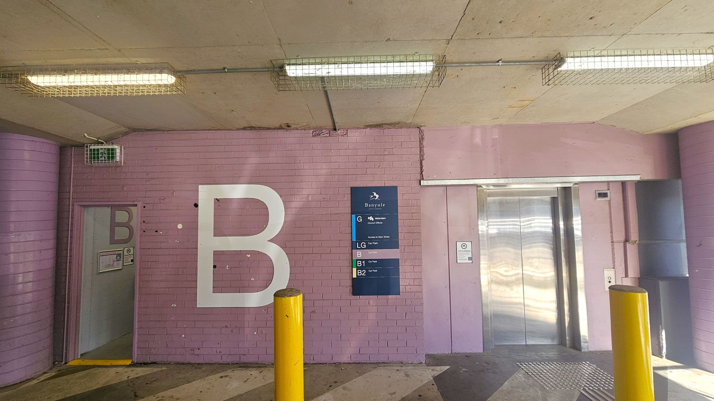 A pink brick wall with a large white letter "B" on the left. A small door with another "B" is partially open. In the centre, a blue sign lists different levels of a building. On the right, there is a metallic lift with closed doors. Two yellow bollards are in front of the wall. The ceiling is concrete with fluorescent lights encased in wire cages. The floor has a striped pattern.