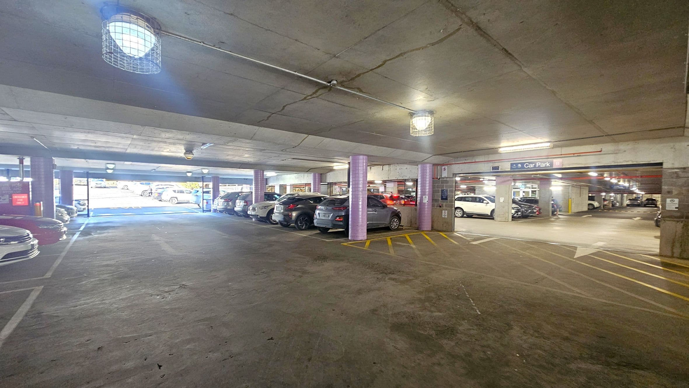 A multi-storey car park with concrete floors and ceilings. On the left, several parked cars are visible, with a bright light coming from the entrance. The right side shows more parked cars and a sign indicating "Car Park B". The ceiling has exposed lighting fixtures with protective cages. The floor has yellow markings and arrows guiding traffic flow. The area is well-lit with natural light from the entrance and artificial lights overhead.