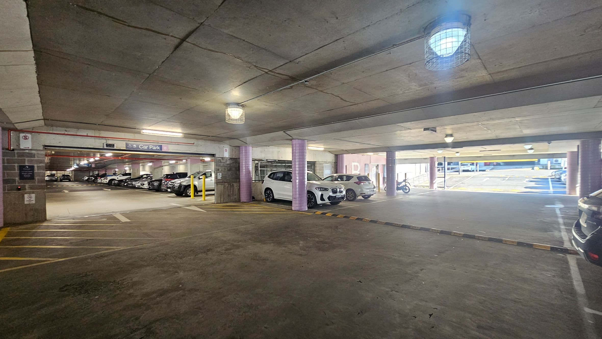 A multi-storey car park with a concrete ceiling and floor. On the left, several parked cars are lined up, with a sign indicating "Car Park B". In the centre, a white car is parked near a purple pillar. On the right, there is an open area leading to an outdoor section with more parking spaces and a motorcycle. The lighting is provided by ceiling fixtures with protective cages. Yellow lines and barriers are visible on the floor.