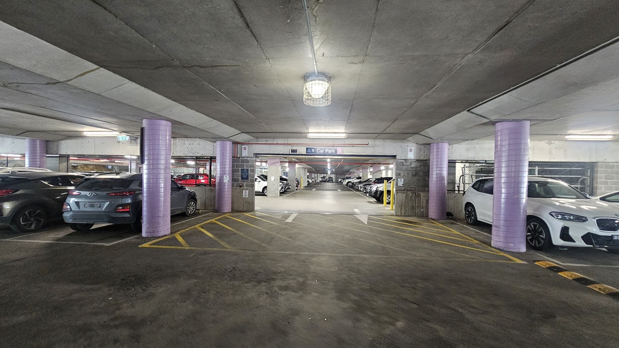 A multi-storey car park with a concrete ceiling and floor. In the foreground, there are yellow diagonal lines on the ground. The middle section shows a wide entrance to another parking area, with a sign above indicating "Car Park." The left side has parked cars, including a grey Hyundai, and the right side features a white car. Purple cylindrical columns are evenly spaced along the entrance. The background reveals more parked cars and fluorescent lighting.