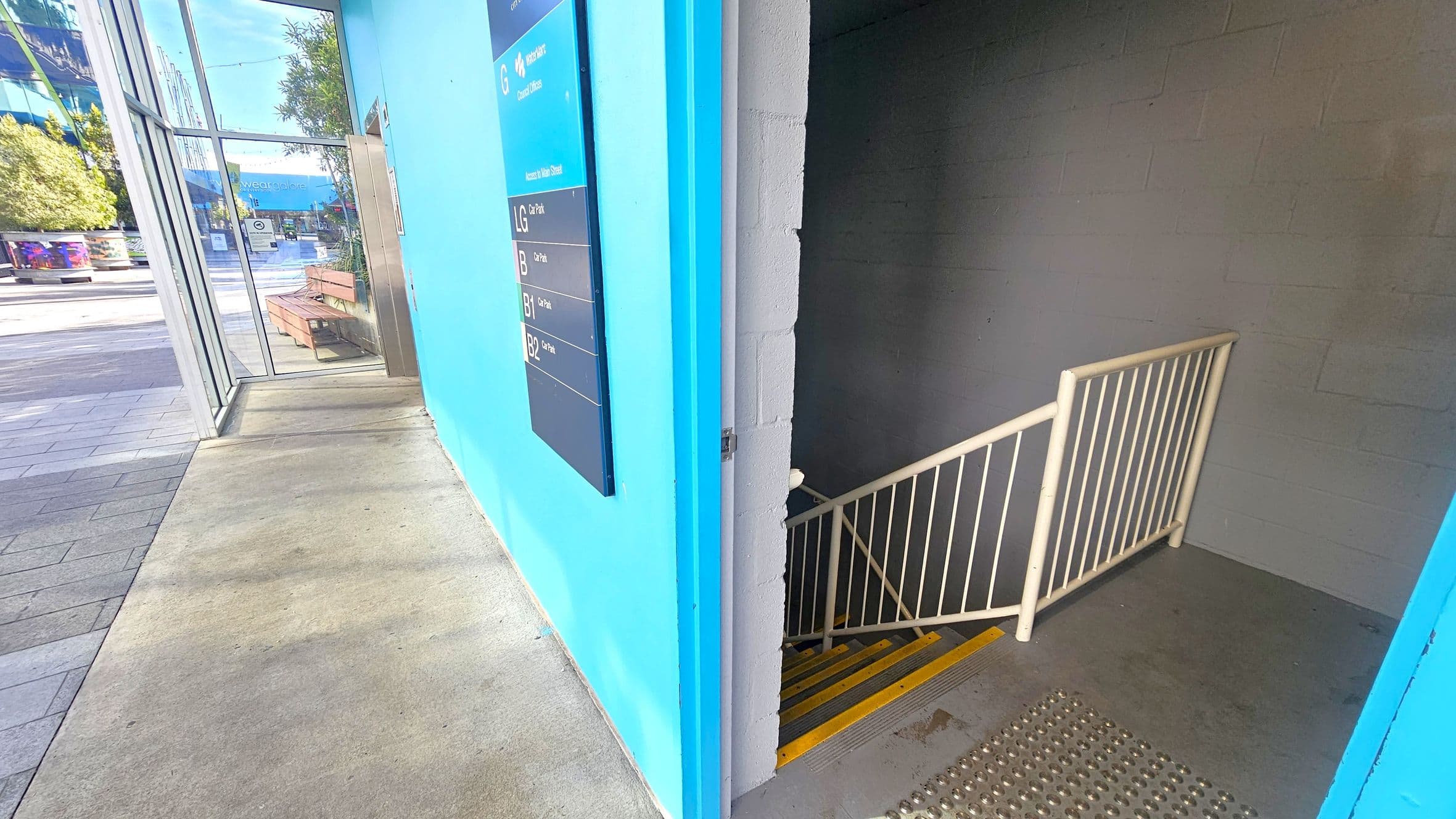 On the left, there's a glass entrance with a view of an outdoor area featuring benches and trees. The floor is a mix of concrete and tiles. On the right, a stairwell with a metal railing descends, marked by yellow safety strips. The wall is painted bright blue with a directory sign listing different levels. The ceiling is not visible.