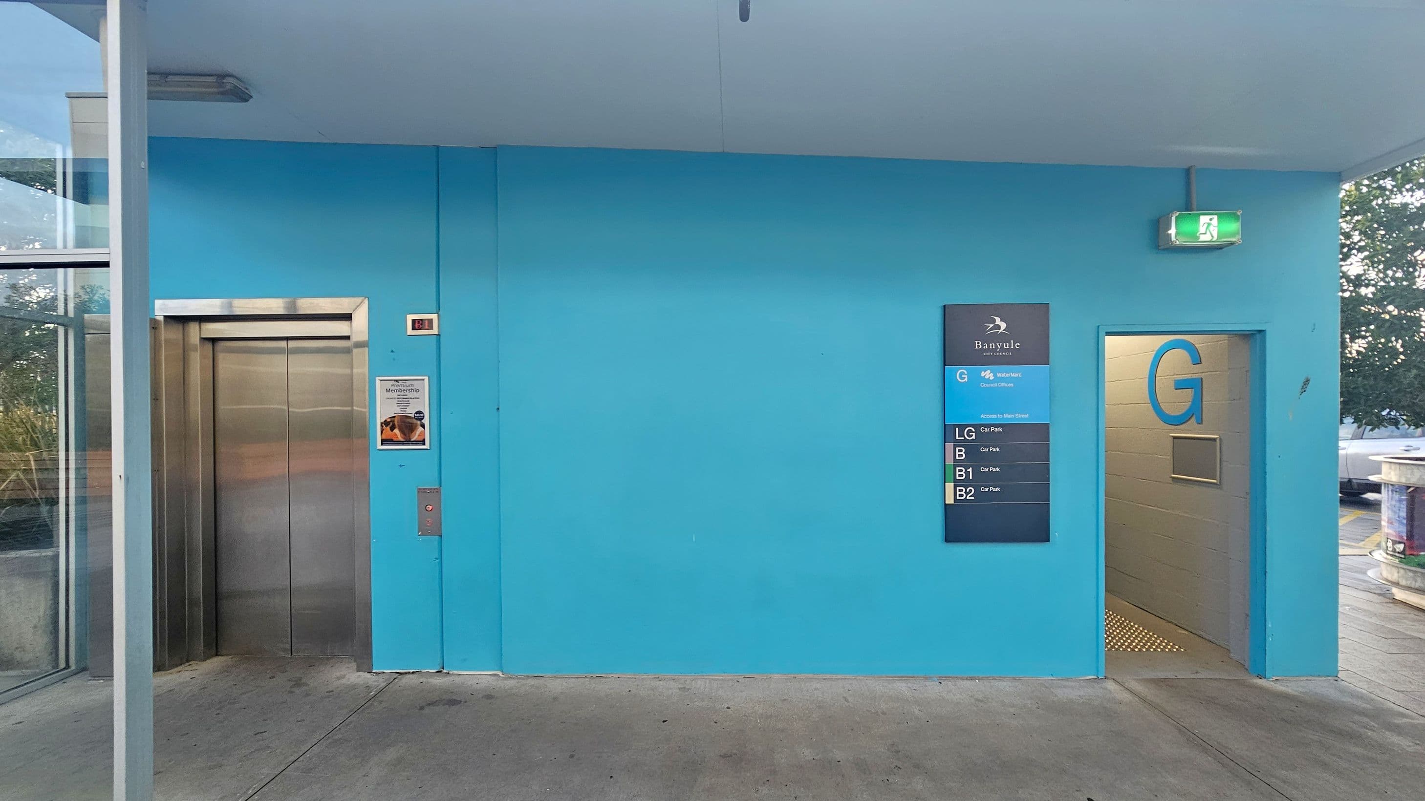 A blue wall with a lift on the left, featuring a stainless steel door and a poster beside it. To the right, a sign displays floor information for a building, labelled "Banyule" with levels G, LG, B, B1, and B2. An open doorway marked "G" leads to a corridor. Above the doorway, a green emergency exit sign is visible. The floor is concrete, and the ceiling is plain. A glass panel on the far left reveals some greenery outside.