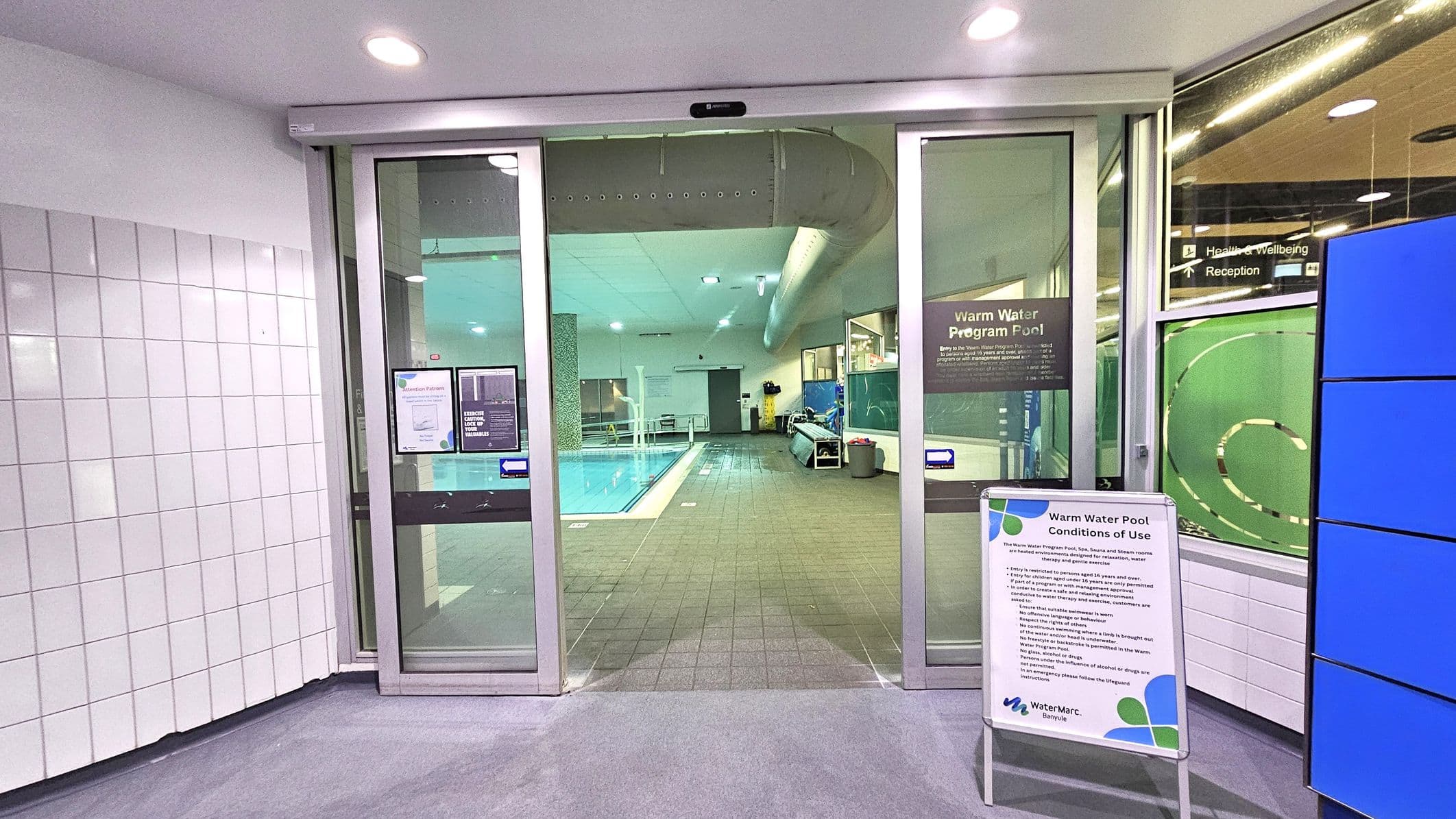 Sliding glass doors lead to an indoor pool area. The pool is visible in the centre, with a tiled floor surrounding it. On the left, white tiled walls are present. On the right, a sign with "Warm Water Pool Conditions of Use" is displayed. The ceiling has recessed lighting, and a large ventilation duct runs along it. In the background, pool equipment and a bench are visible.
