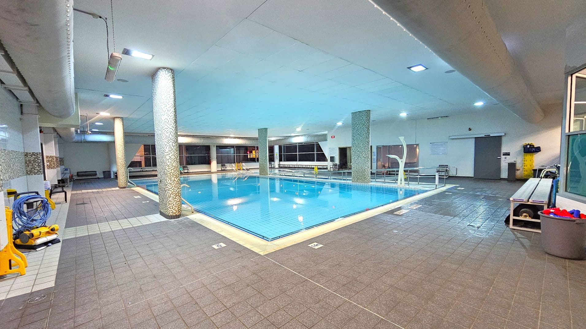 An indoor swimming pool with clear blue water is surrounded by a tiled floor. On the left, there are cleaning equipment and a coiled hose. The ceiling features recessed lighting and large ventilation ducts. In the background, large windows line the walls, and there are several benches and a door. A bin with red and blue items is on the right, next to a bench. The pool is bordered by mosaic-tiled columns.