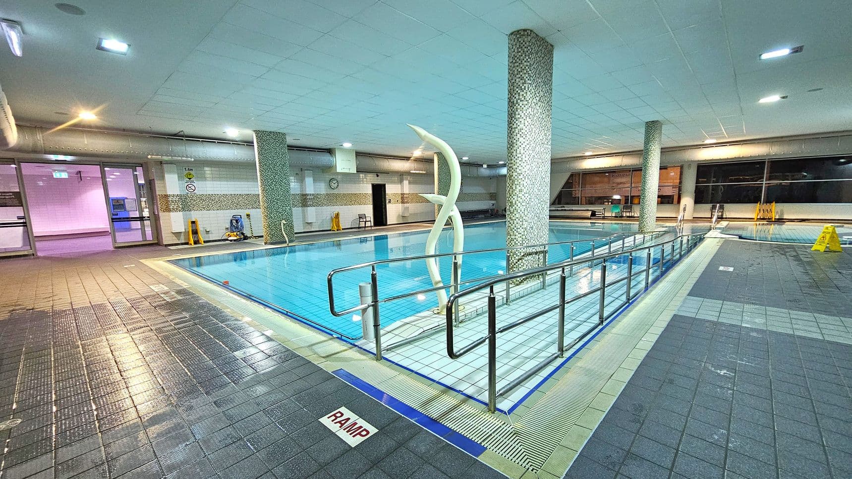 An indoor swimming pool with a tiled floor and ceiling. In the centre, a pool with clear blue water is surrounded by a metal railing. A decorative sculpture stands near a mosaic-tiled column. On the left, glass doors lead to another area, and a clock is visible on the wall. On the right, large windows and a few caution signs are present. The floor is wet, with a marked ramp area in the foreground. The lighting is bright and evenly distributed.