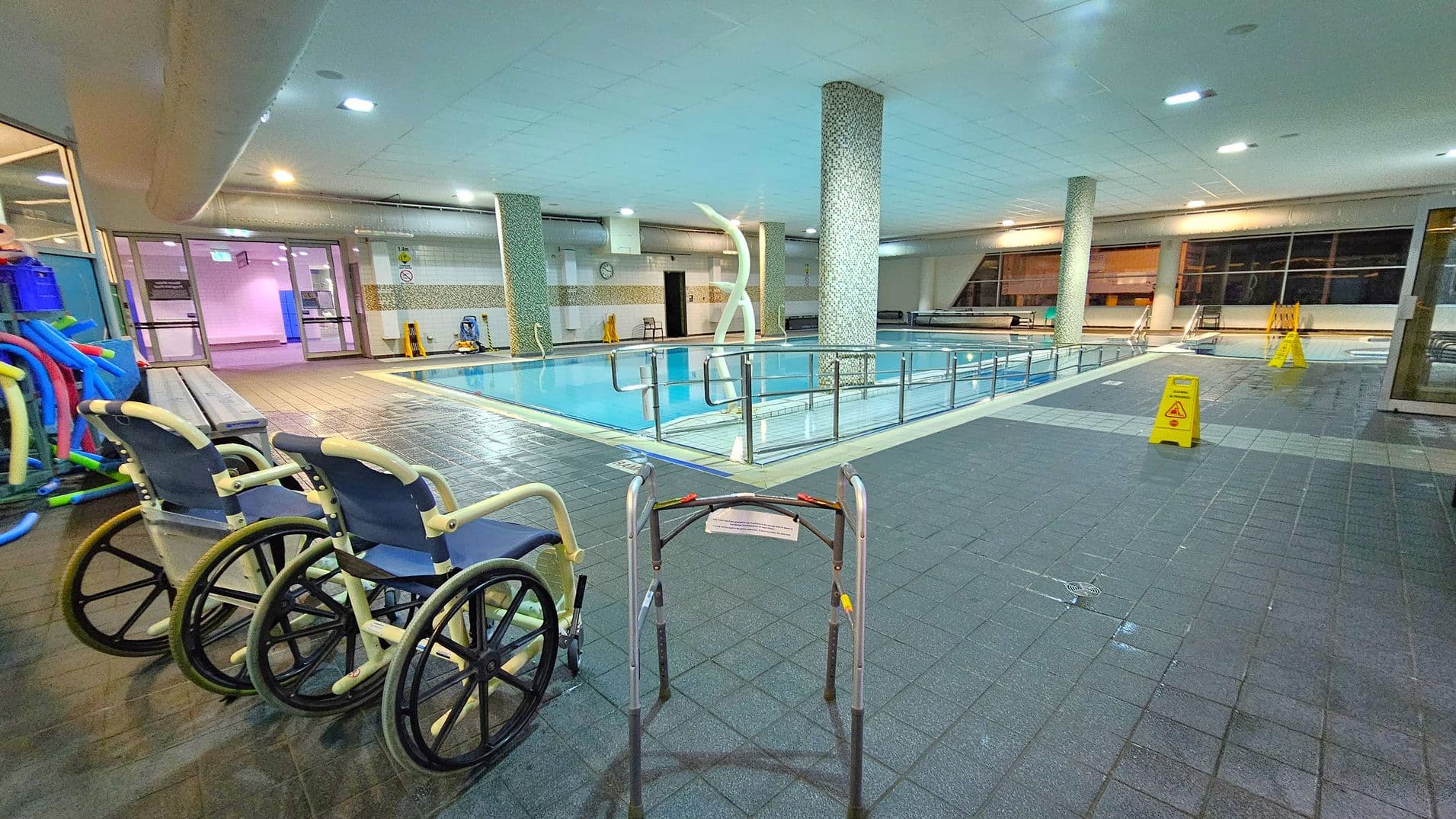 An indoor swimming pool area with a tiled floor and ceiling. On the left, there are two wheelchairs and a collection of pool noodles. In the centre, a pool is surrounded by a metal railing, with a decorative sculpture nearby. On the right, there are caution signs on the floor. Large windows line the background, and there are benches along the walls. The area is well-lit with overhead lights.