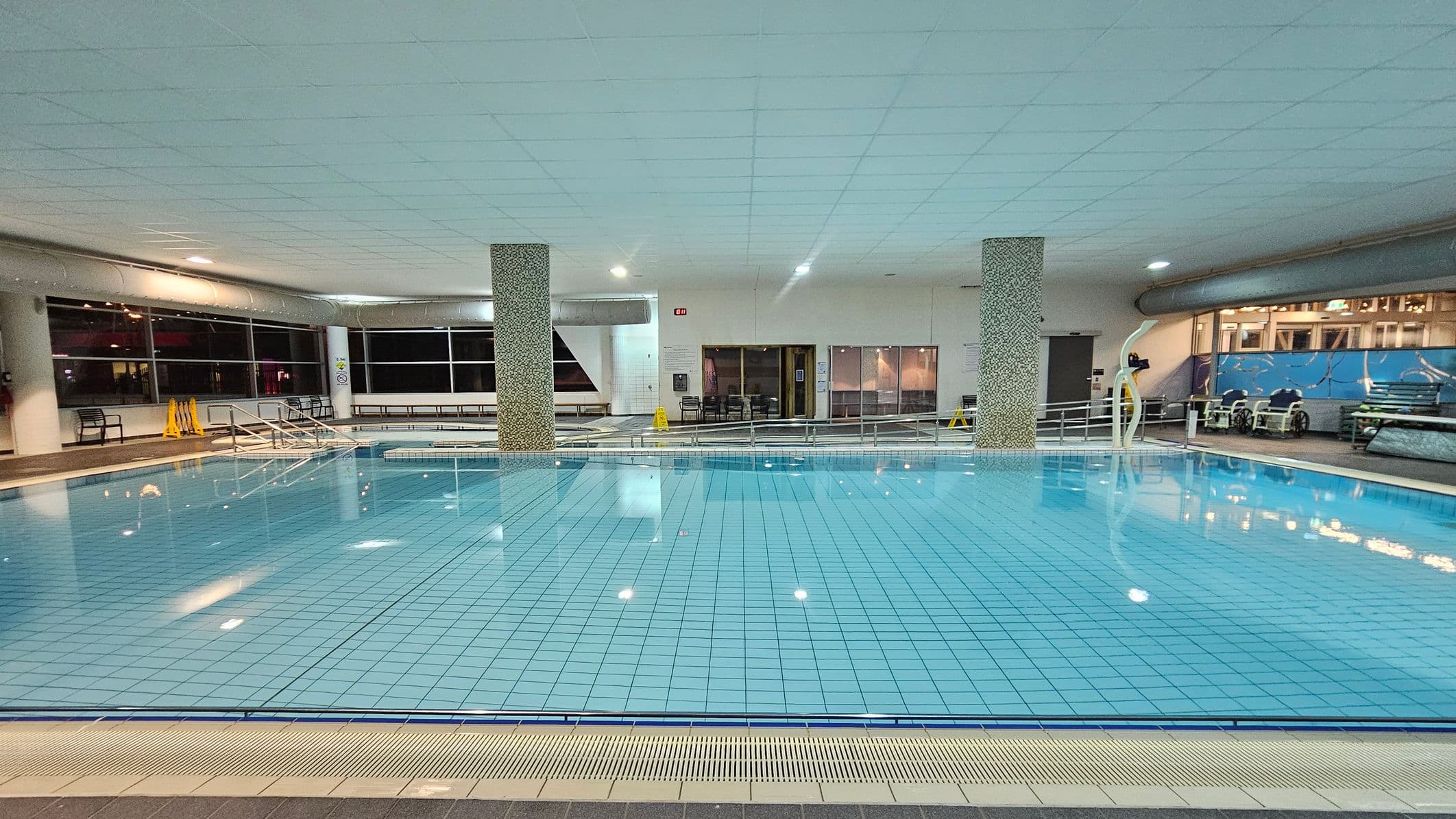 Indoor swimming pool with clear blue water and tiled floor. Two mosaic-covered pillars are in the centre. On the left, large windows and a few benches are visible, with yellow caution signs nearby. The right side has a mural and some poolside equipment. The ceiling is white with recessed lighting, and the pool area is surrounded by a tiled walkway.