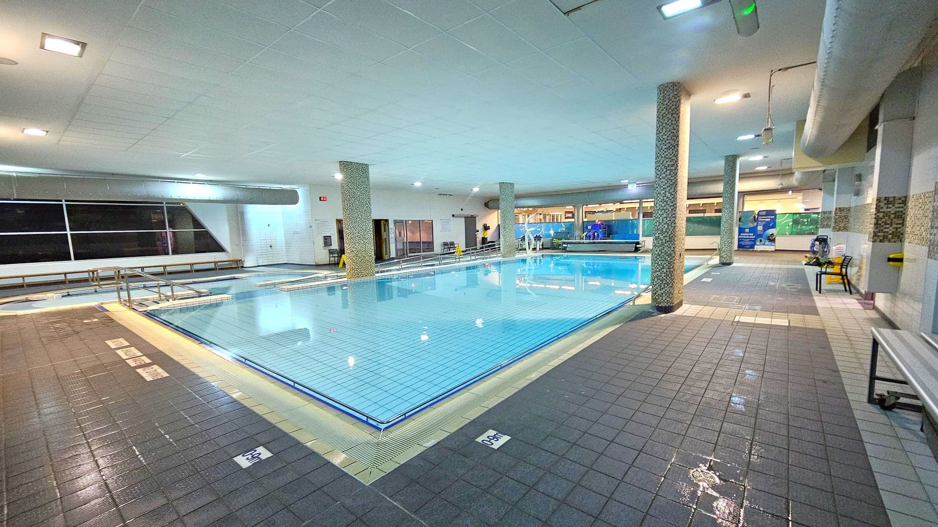 An indoor swimming pool with clear blue water is surrounded by a tiled deck. On the left, there are large windows and a row of benches. In the centre, two mosaic-tiled columns support the ceiling. On the right, there are informational posters and a yellow chair. The ceiling is white with recessed lighting, and the floor is covered with dark grey tiles, some of which are wet. A metal handrail leads into the pool.