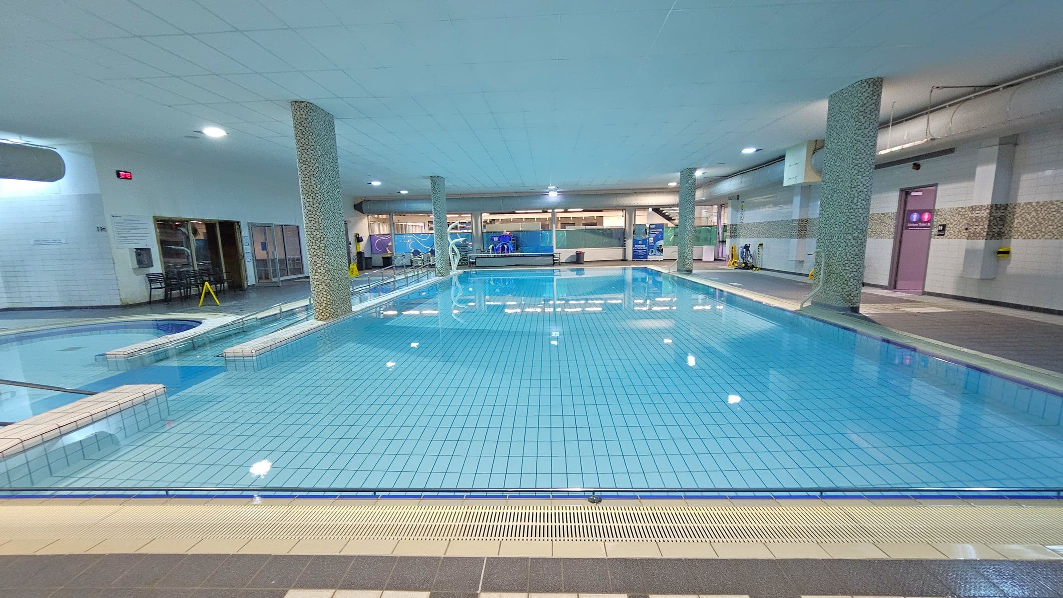 An indoor swimming pool with clear blue water and tiled flooring. Several mosaic-tiled columns are positioned around the pool. On the left, there is a smaller, circular pool area. The background features large windows and doors, with a staircase visible on the right. The ceiling is white with recessed lighting, and the floor surrounding the pool is tiled in shades of grey and beige. A caution sign is placed near the entrance on the left.