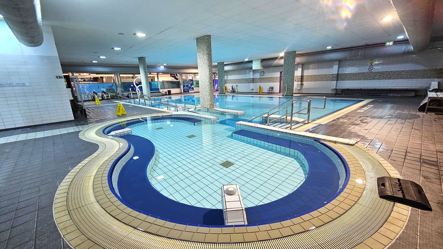 An indoor swimming pool area with a curvy, shallow pool in the foreground, surrounded by a beige and blue tiled border. The main pool is in the background, featuring handrails and a tiled deck. Several columns support the ceiling, which is fitted with recessed lighting. The floor is tiled in shades of grey and beige, with caution signs visible near the pool. Benches and a wall with a clock are on the right side.