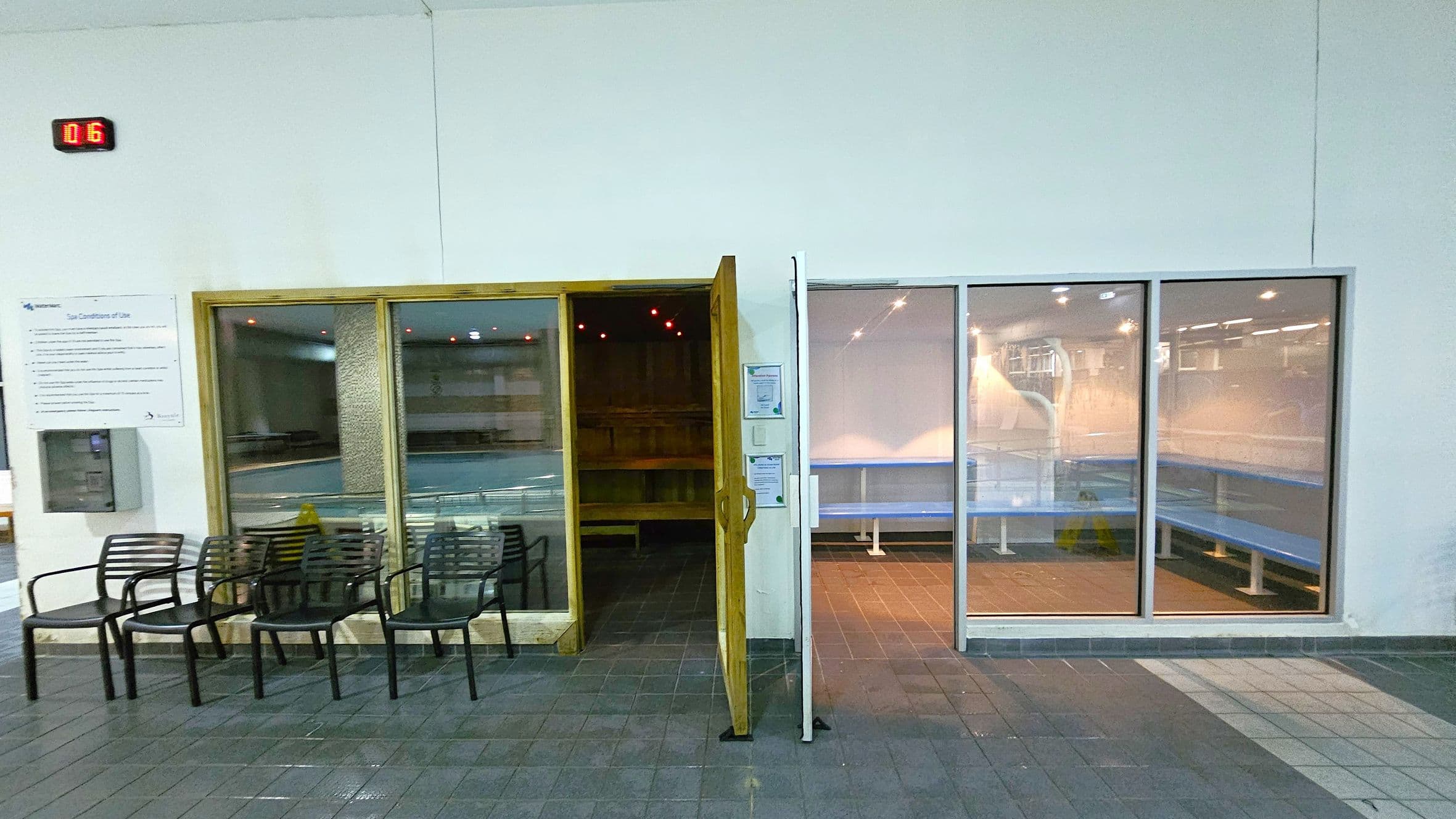 On the left, there are several black chairs lined up against a white wall. Above them, a digital display shows "016". A sign with text is mounted on the wall. In the centre, a wooden-framed glass door is open, revealing a dimly lit sauna with wooden benches. On the right, large glass panels show a room with blue benches and bright lighting. The floor is tiled in shades of grey and green.