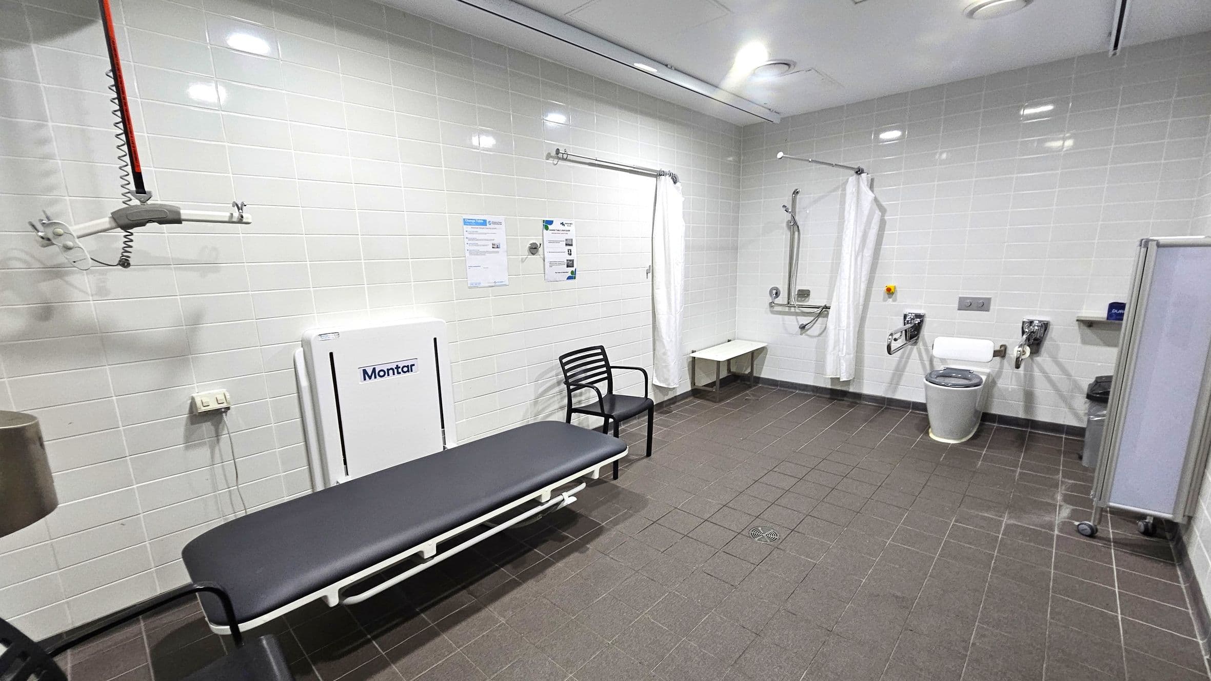 A spacious, accessible bathroom with white tiled walls and a dark tiled floor. On the left, there's a black bench with a white frame, a black chair, and a wall-mounted hoist system. In the centre, a white curtain hangs from a rail. On the right, there's a toilet with support bars, a small shelf, and a bin. The ceiling is white with recessed lighting. A foldable changing table is mounted on the wall.