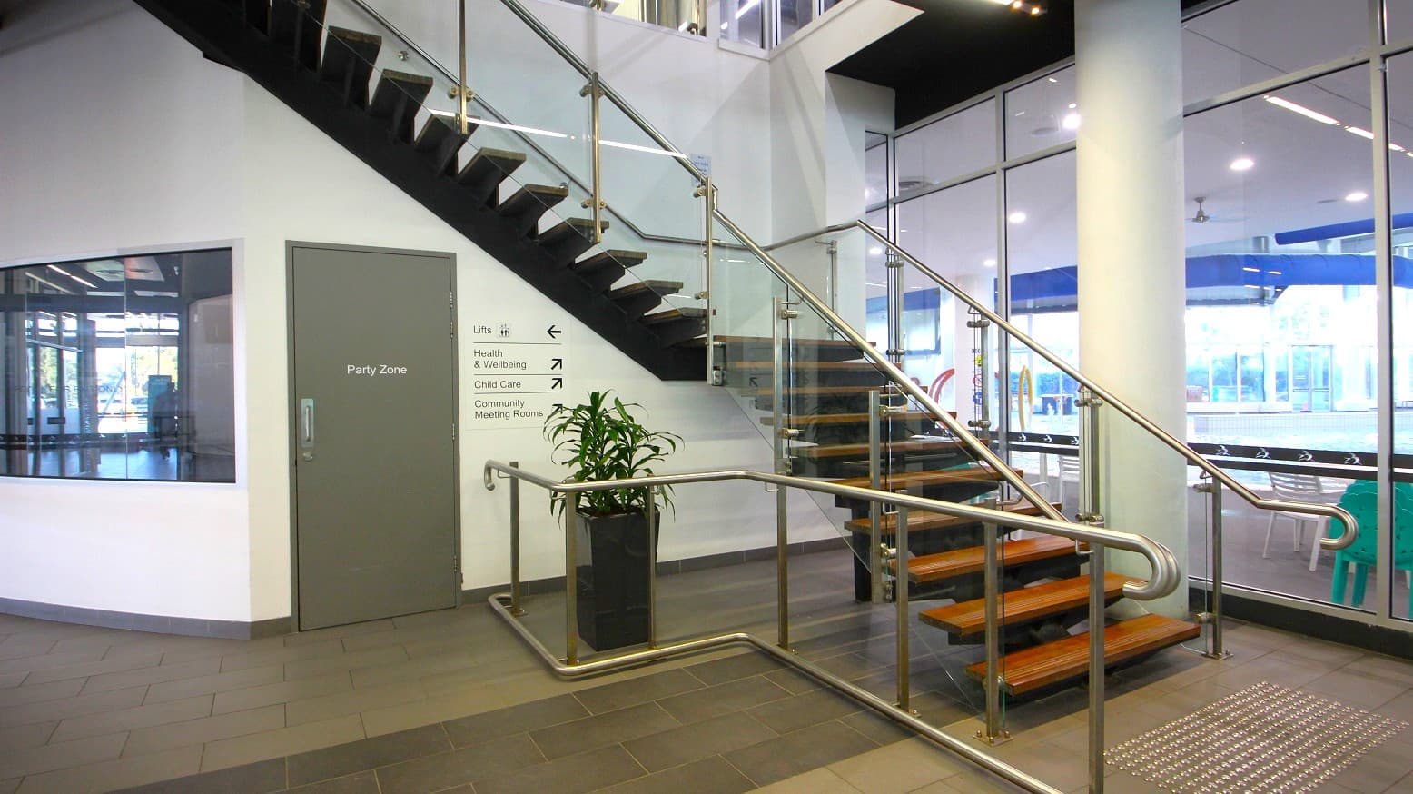 A modern indoor space with a staircase featuring wooden steps and glass railings. On the left, there's a grey door labelled "Party Zone" and a wall with directional signs. A potted plant sits near the base of the stairs. The floor is tiled, and large glass windows on the right reveal a view of an outdoor area with tables and chairs. The ceiling has recessed lighting.