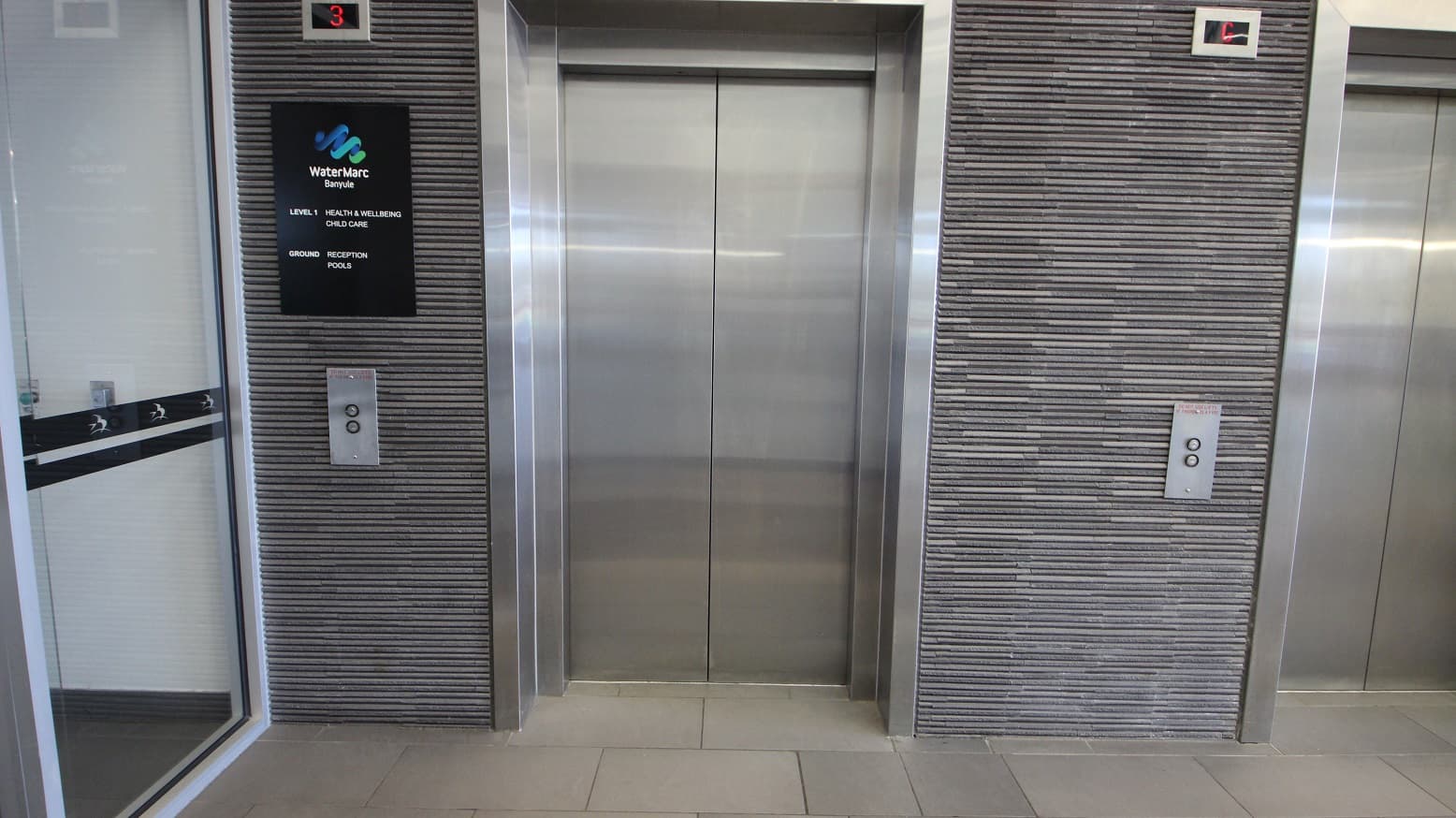 Two lift doors are visible, with the one on the left partially open, revealing a mirrored interior. The central lift door is closed, with a metallic finish. Both lifts are set within a wall covered in horizontal grey tiles. Above the left lift is a sign for "WaterMarc Banyule" with floor information. Each lift has a control panel with buttons. The floor is tiled in light grey, and there is a small display above each lift showing the floor number.
