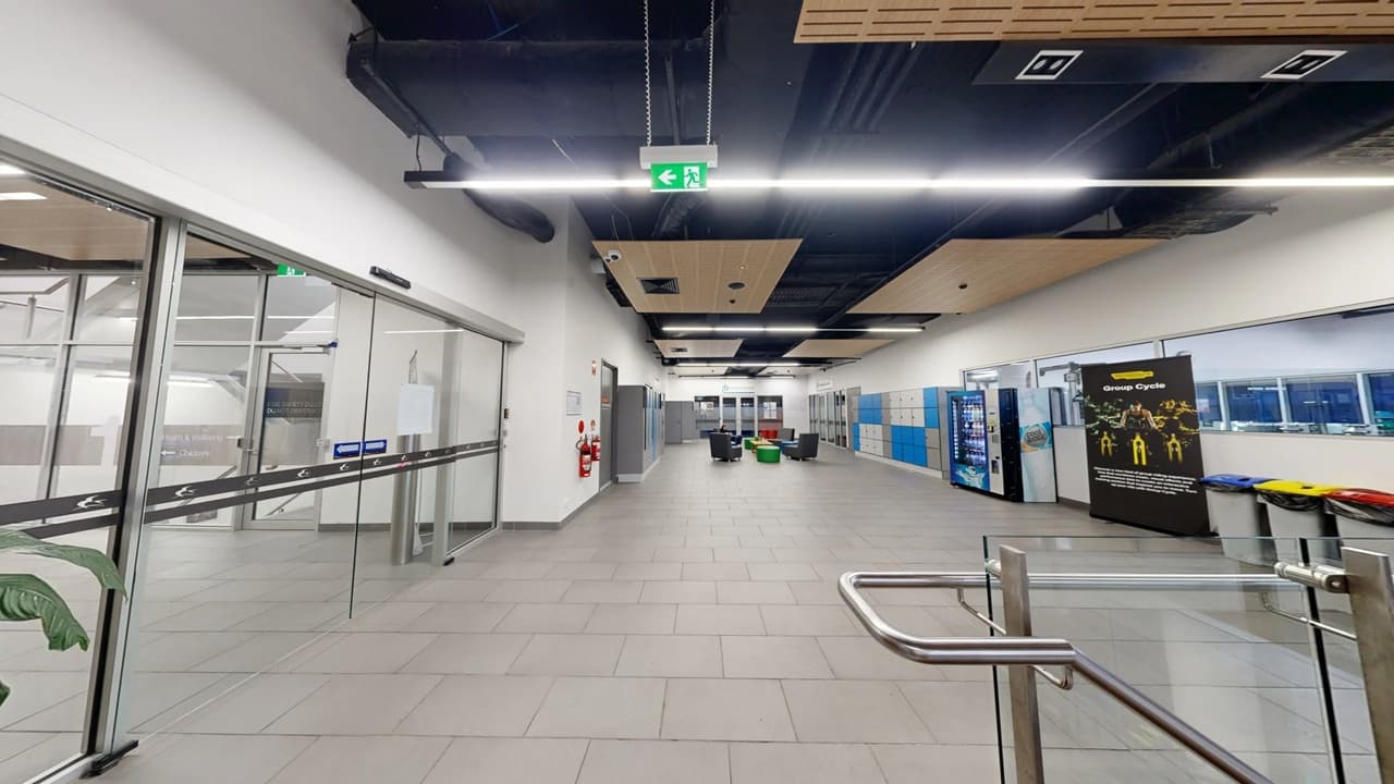 A spacious indoor area with a grey tiled floor and a ceiling featuring black and wooden panels. On the left, there are large glass doors with a plant visible through them. In the centre, there are seating arrangements with chairs and tables. On the right, vending machines and a display board are present, along with a row of colourful bins. An exit sign hangs from the ceiling, and a metal barrier is in the foreground.