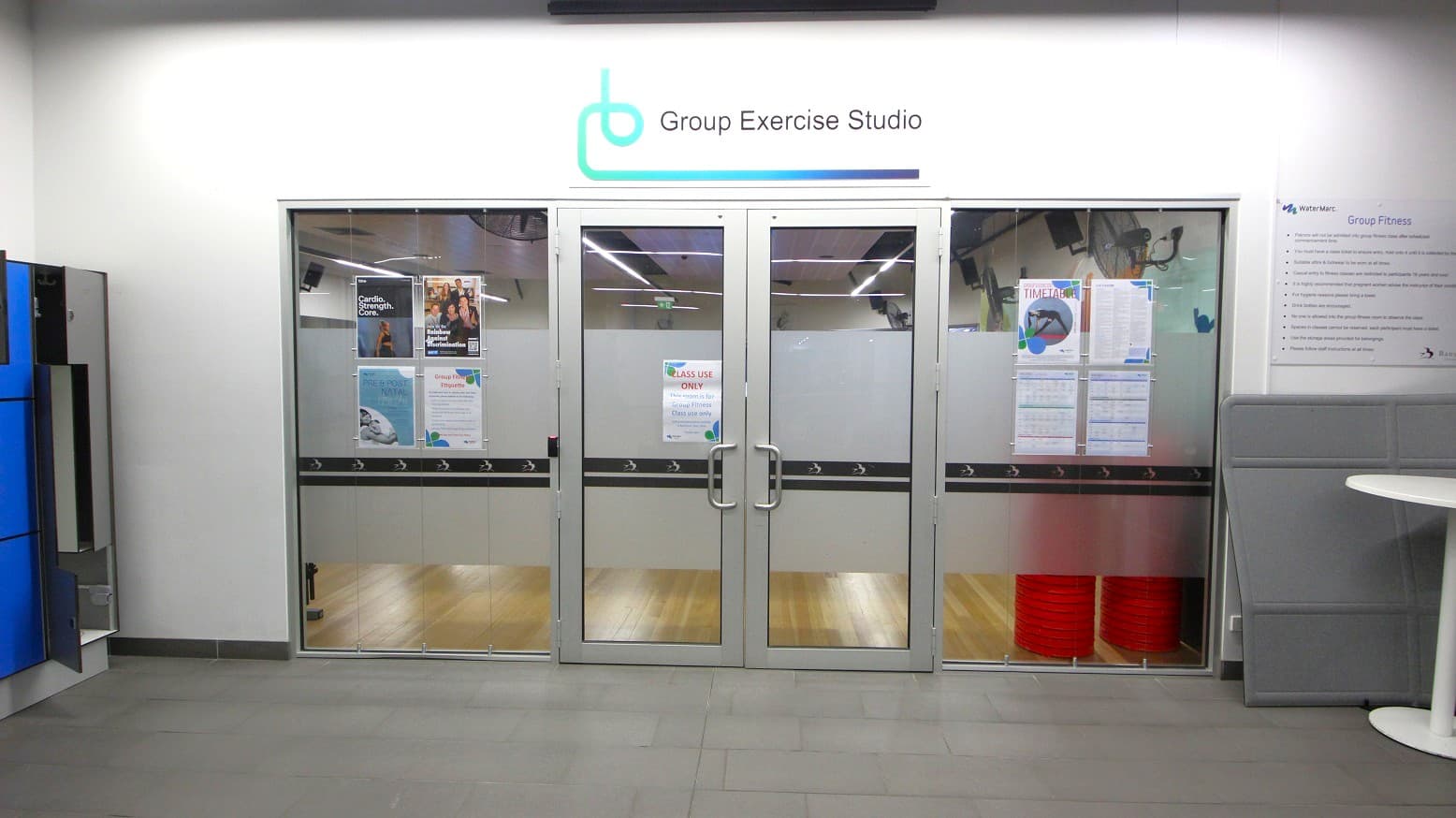 Glass doors leading to a group exercise studio, with posters and notices displayed on them. Above the doors, a sign reads "Group Exercise Studio" with a logo. The floor inside is wooden, while the exterior floor is tiled. On the left, there are blue lockers, and on the right, a white round table and a grey partition. The ceiling has fluorescent lights.