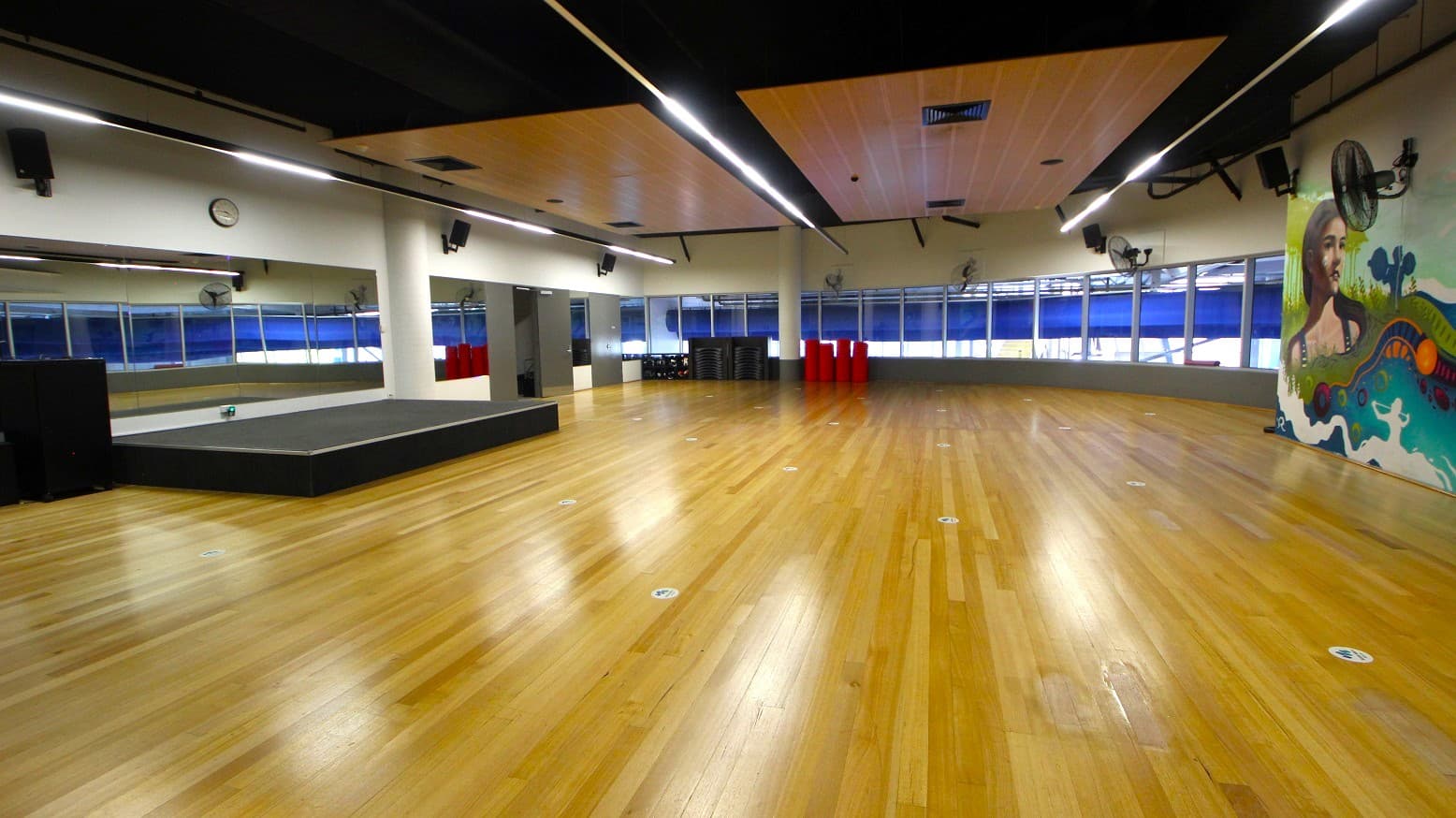 A spacious room with a polished wooden floor and a high ceiling featuring recessed lighting. On the left, there's a large mirror and a small stage. The back wall has large windows with blue blinds. On the right, a colourful mural is visible. Several red cylindrical objects and stacked black equipment are against the back wall. Ceiling fans are mounted on the walls.
