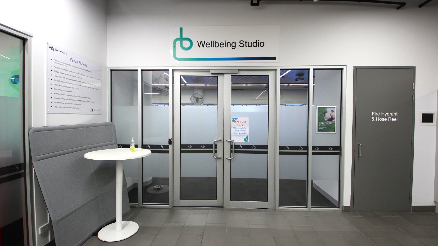 Entrance to a "Wellbeing Studio" with glass double doors in the centre, featuring a sign above. To the right, a grey door marked "Fire Hydrant & Hose Reel". On the left, a white round table with a small bottle on top, next to a grey padded panel. A sign on the wall lists "Group Fitness" information. The floor is tiled in grey, and the ceiling has visible lighting fixtures.