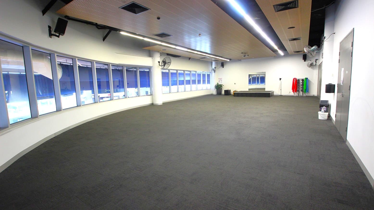 A spacious room with a curved wall of large windows on the left, allowing natural light to enter. The floor is covered with grey carpet. On the right, there is a wall with a door and some cleaning supplies. At the far end, there are exercise mats in various colours stacked vertically, a small platform, and a potted plant. The ceiling features recessed lighting and ventilation ducts.