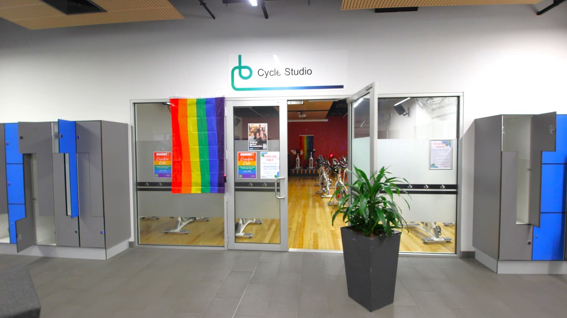 A cycle studio entrance with glass doors, displaying a rainbow flag on the left door. Above the doors, a sign reads "Cycle Studio". Inside, several stationary bikes are visible on a wooden floor. On the left and right of the entrance, there are grey lockers with blue doors, some of which are open. A large potted plant is positioned to the right of the entrance. The ceiling has wooden panels, and the floor is grey.