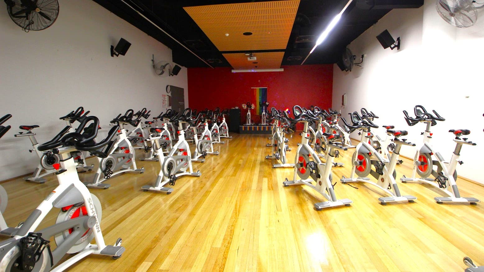 A spacious indoor cycling studio with numerous stationary bikes arranged in rows on a polished wooden floor. The walls are white, with a vibrant red accent wall at the back. Ceiling fans and speakers are mounted on the side walls. The ceiling features a wooden panel section in the centre. At the back, there is a small stage with additional bikes and a colourful vertical banner. The room is well-lit, creating a bright atmosphere.