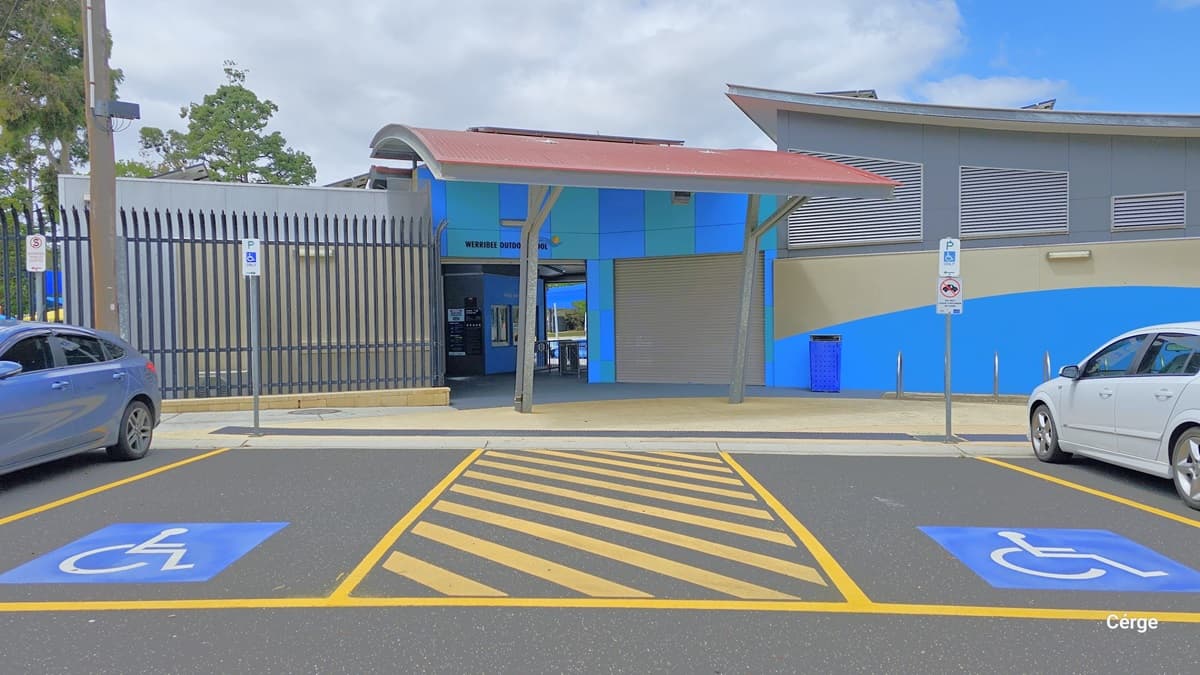 Two accessible car parking spaces positioned directly outside the entrance of Werribee Outdoor Pool. Each accessible car parking space has a white wheelchair logo for persons with mobility disabilities over a blue background and is separated by yellow rectangular line markings filled with diagonal yellow lines inside. Two parking signs for Persons with mobility disabilities and Do not leave children in cabs are placed within the car park spaces.