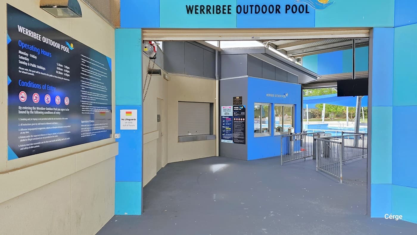 Sky blue and light blue entrance arch with a rectangular tile design at Werribee Outdoor Pool. On the top of this arch is the name of the facility, detailed in black typeface with the logo containing the yellow-orange element for the sun above the blue wave element for water. On the left cream wall is a large blue signboard for the operating hours, conditions of entry, and supervision of children. The entrance has grey floors that lead to the reception window and tripod turnstile gate.