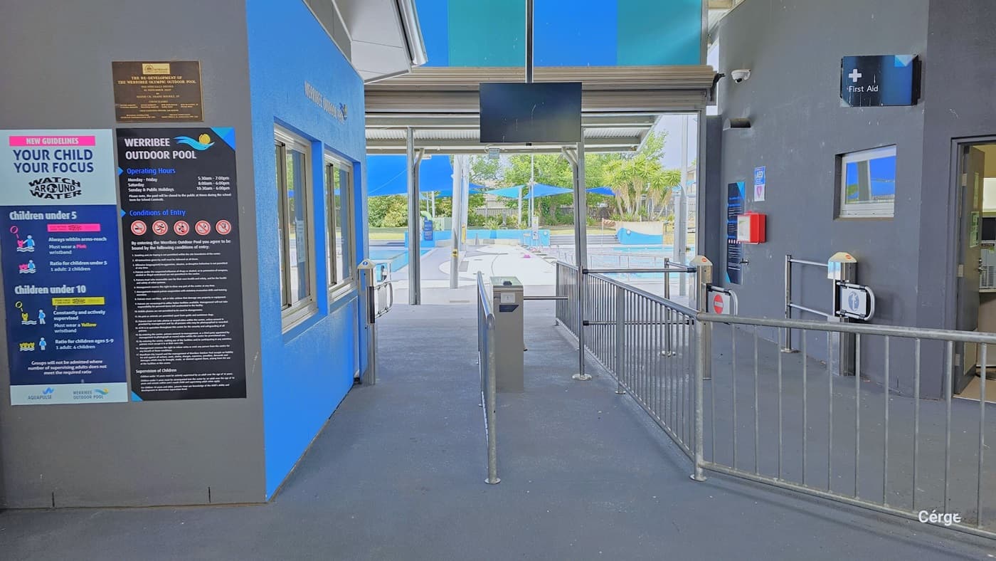 Two sets of turnstile gates for the entry and exit points of the facility are separated by a grey low-set barrier fence. A black monitor screen overhangs in the middle of the pathway. On the grey back wall of the reception room are smaller, elongated rectangular versions of the conditions of entry and the colorful infographic for the supervision of children under 5 and under 10. On the light blue reception wall are two sets of glass windows with white metal frames.