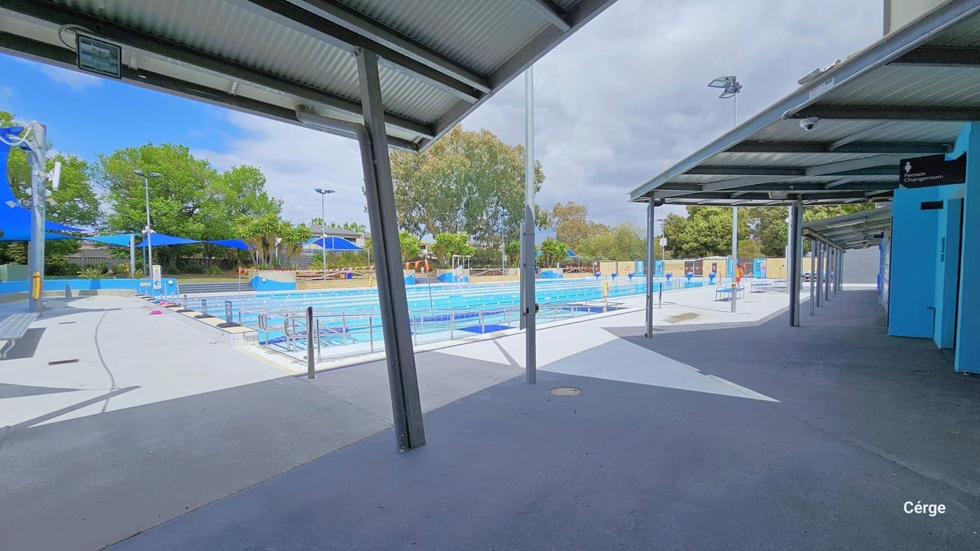 Overlooking view of the 50m, eight-lane Werribee outdoor pool. The pool has ramp access with steel handrails for people with mobility disabilities. Near the admin building is a floor painted in grey. On the far right is the Female Change Room sign placed on the upper part of the sky-blue wall. On one of the metal beams is a CCTV camera facing the entrance of the female change room.
