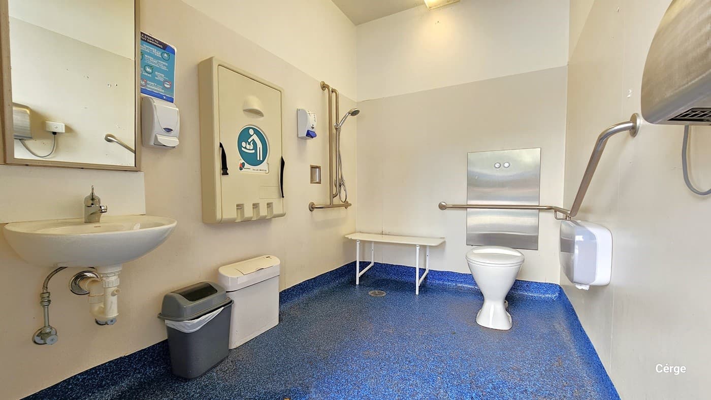 Another accessible bathroom at Werribee Outdoor Pool. The bathroom has a blue stonelike tile surface and a half-cream and white wall. Mounted on the left wall are the shower head, a cream baby change table, a liquid soap dispenser, a mirror, and a wash basin. There is also a grey garbage bin with a white garbage bag. Behind the toilet is a grab bar, while to the left is a white shower bench. Mounted on the right wall is the stainless steel hand dryer and a white and grey tissue holder.