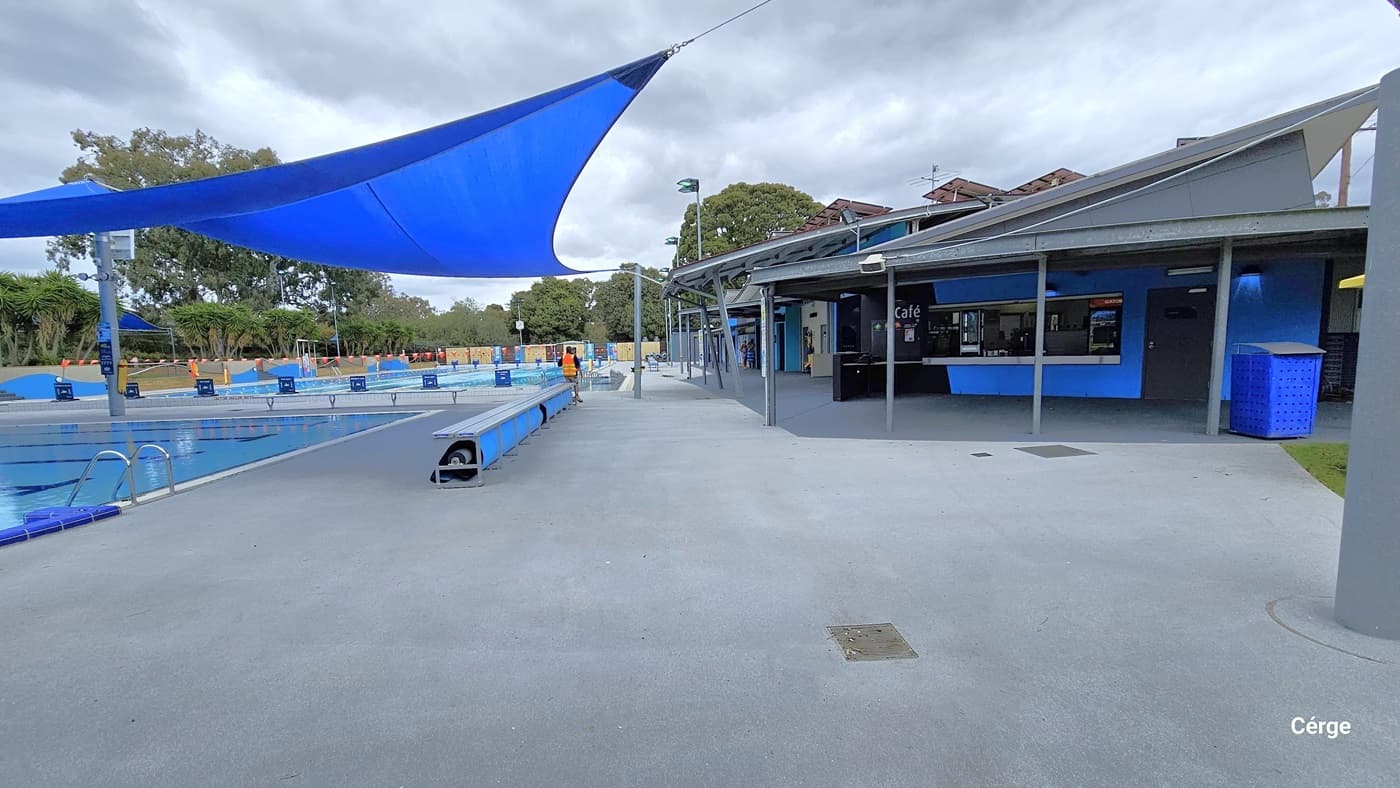 View near the heated leisure pool, near the grey metal bench with a blue sunshade overhang that protects it from overexposure to sunlight. Further to the left is the 50m, eight-lane pool with blue signs on its pool deck at one end and strings of orange rectangular flags. To the right is the café. The image also shows the unique architectural roofing of the café, which has an inclining roof style down the pool with a circular roof pattern underneath.