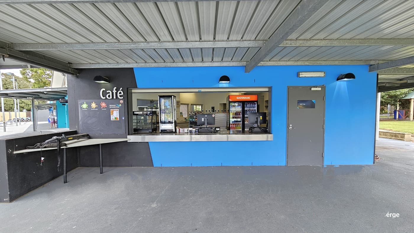 Front view of the café at Werribee Outdoor Pool. The café has an overall scheme of grey and blue. There is a white triangular table with black metal legs on the left mid-high wall and in front of the black wall with a bulletin board. Seen through the open countertop are the snacks, hot meals, and the fridge for the cold refreshments. The ceiling is made of grey corrugated metal beams, and the floor is grey. Black small LED wall lamps are placed on the upper part of the wall.
