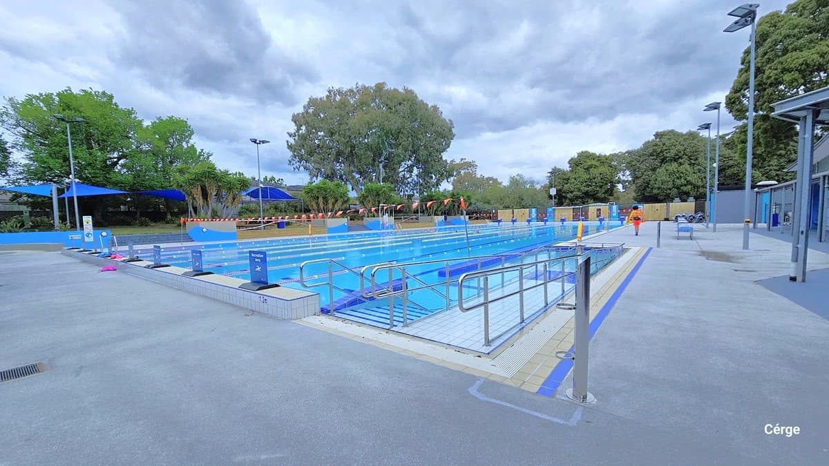 Ramp and stair access with handrails of the eight-lane, 50m pool. There is a small opening after the ramp and stairs that is created by the blue-tiled pool walls. At the top of the white tile pool coping are blue signboards with flat black floor supports. Each lane is separated by floating rope lanes. Strings of orange triangular flags are around the opposite poles of the pool. Further to the back are brown walls, while to the left are blue sunshades.